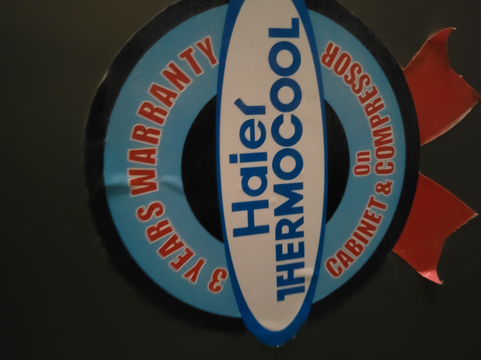 The image shows a close-up of a sticker or emblem related to a refrigerator. The sticker is circular, with a light blue ring around a black center. The words "3 YEARS WARRANTY" are printed in red on the upper left of the blue ring, and "CABINET & COMPRESSOR ON" are printed in red on the lower right of the blue ring. A white, elongated oval shape is superimposed over the center, with the words "Haier THERMOCOOL" printed vertically in blue on the oval. Two red, flag-like shapes are attached to the right side of the sticker. The background is dark, with some glare visible on the sticker. The city and location are unknown.