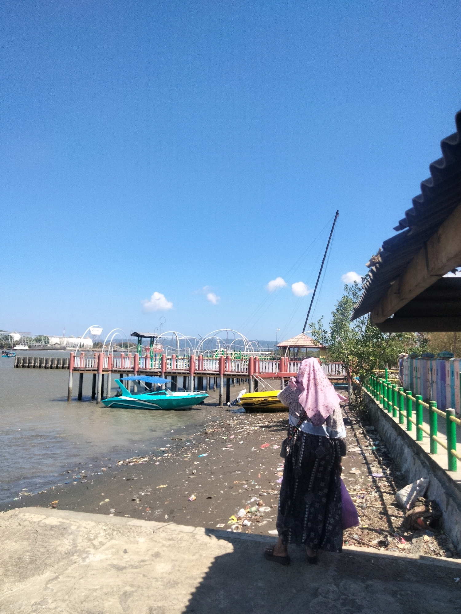 A wide shot captures a sunny day by a body of water in Cilacap, Indonesia. The sky is a clear, bright blue with a few wispy clouds. In the foreground, a person wearing a pink hijab and a patterned skirt stands on a concrete walkway. The person is facing away from the camera, looking towards a wooden pier that extends into the water. The pier has a red and white railing and is lined with decorative white arches.
Several boats are visible. A bright teal and white speedboat is docked near the shore, partially on the sand. A yellow boat is also visible near the pier. The shoreline is covered in scattered debris, including plastic bottles and wrappers.
Beyond the pier, a long, thin mast of a sailboat stands tall against the sky. In the distance, industrial structures and buildings can be seen along the coastline, suggesting an urban or industrial area. The overall scene depicts a waterfront location, likely a tourist or recreational spot, marred by visible pollution on the beach.