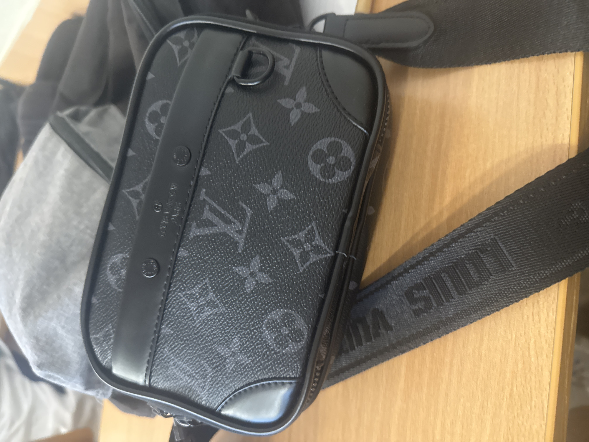 The image displays a close-up, slightly overhead view of a black Louis Vuitton bag placed on a light wooden surface. The main subject is a compact, rectangular-shaped bag, possibly a crossbody pouch, made of black material with a subtle, dark grey Louis Vuitton monogram pattern. It features black trim along its edges and a vertical black strap-like element on the front. On this central strap, faint text reads "LOUIS VUITTON PARIS" alongside a registered trademark symbol. A dark D-ring is visible at the top left corner of the bag.
A black textile strap, presumably attached to the bag, extends across the lower right portion of the frame. This strap features the words "LOUIS VUITTON" repeatedly woven or embossed into its surface in a darker shade of black. The bag rests on a light-toned wooden surface, likely a table or desk. In the blurry background, a light grey object, possibly another bag or piece of fabric, is partially visible. The scene appears to be indoors, and no specific activity is taking place. While the context of Burgas, Bulgaria, is provided, there are no visual cues within the image to confirm this location.