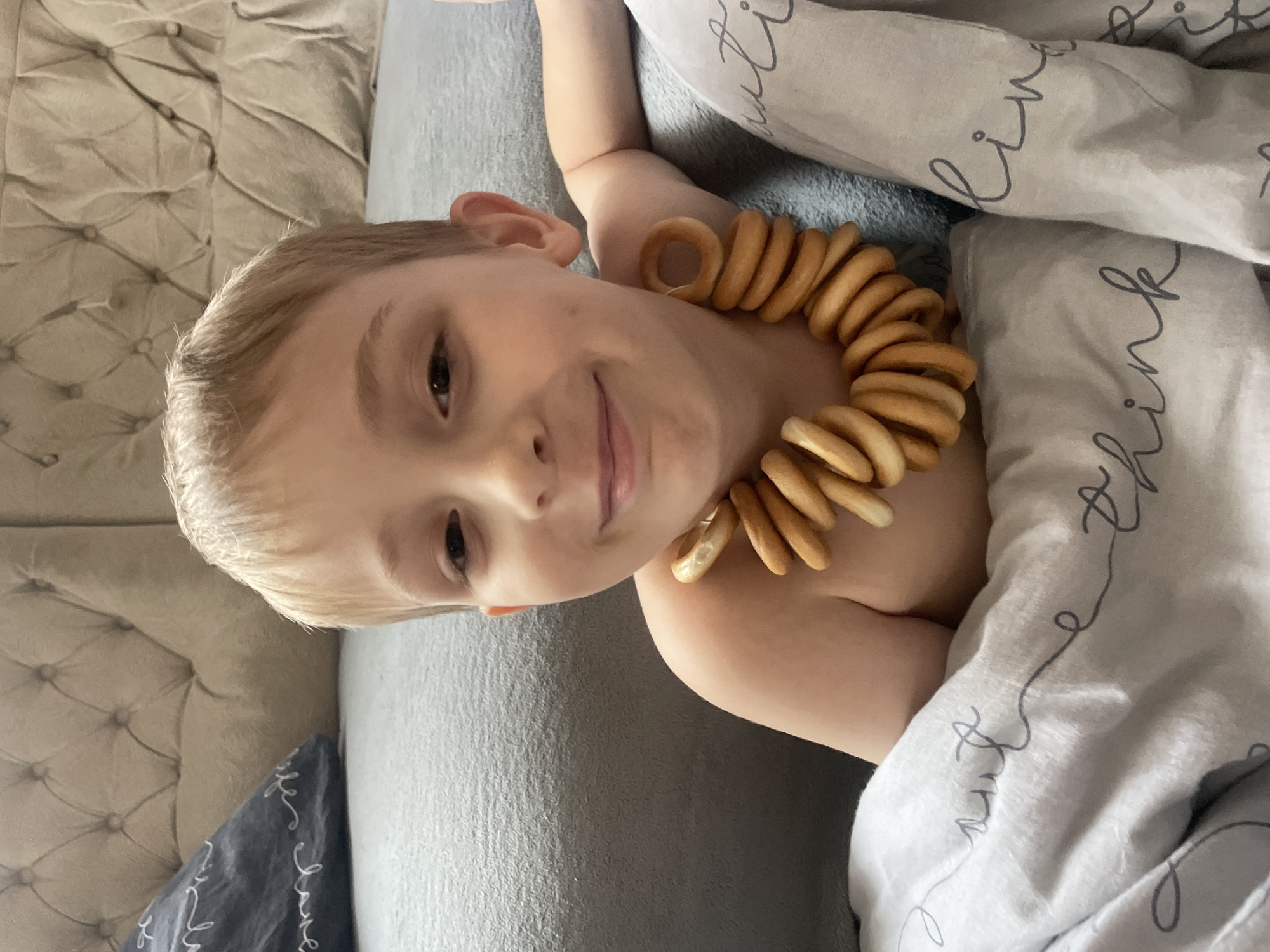 A fair-skinned child with blonde hair is lying on a grey bed, partially covered by white and grey bedding. The child is wearing a necklace made of small, circular, baked goods. The location is unknown, and there is no information to suggest that this image was taken in Baranovka, Russia.