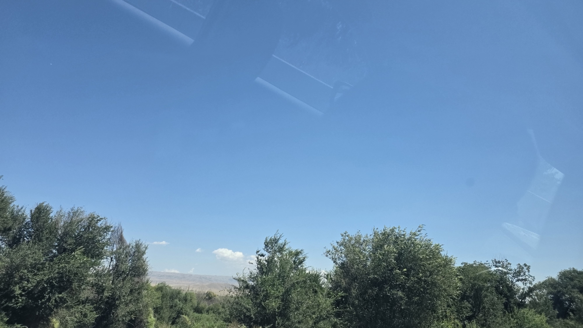 The image shows a clear blue sky with scattered clouds and reflections of objects, likely from a window. In the foreground, a dense line of green trees runs horizontally across the lower part of the frame. Behind the trees, a hilly landscape is visible in the distance, also appearing to be dry and arid. The scene suggests an outdoor environment, possibly during daylight hours. The specific location within the city of Keŋ-Buluŋ, Kyrgyzstan cannot be determined from the visual information provided in the image.