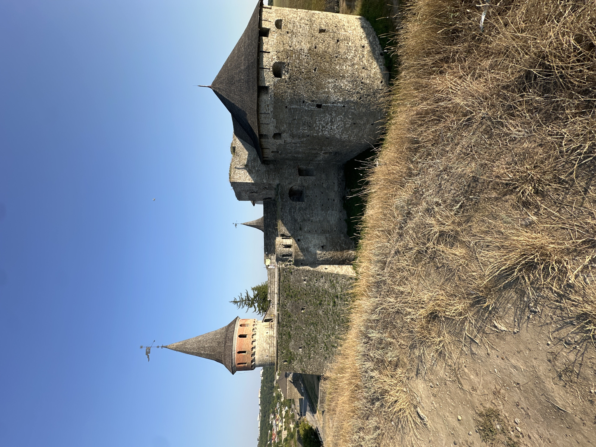 The image shows the Kamianets-Podilskyi Castle in Ukraine, a historical fortress situated on a rocky island, fortified by the Smotrych River canyon. The castle, constructed from grey stone, features several prominent towers with conical roofs. The foreground is dominated by dry, tall grass and sandy soil. The sky above is clear and blue. The castle appears to be an ancient, well-preserved structure.