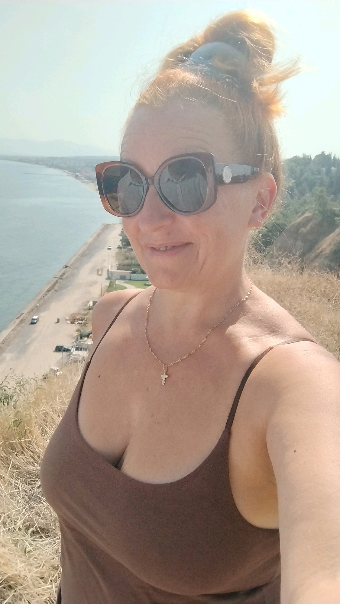 A woman with auburn hair in a messy bun, wearing large brown sunglasses, a brown tank top, and a gold necklace with a cross pendant, takes a selfie outdoors. She is standing on a grassy, dry hillside overlooking a body of water and a coastal road. The background shows a long stretch of coastline with buildings, cars, and hills. The sky is bright and hazy. The location is Agia Triada, Greece.