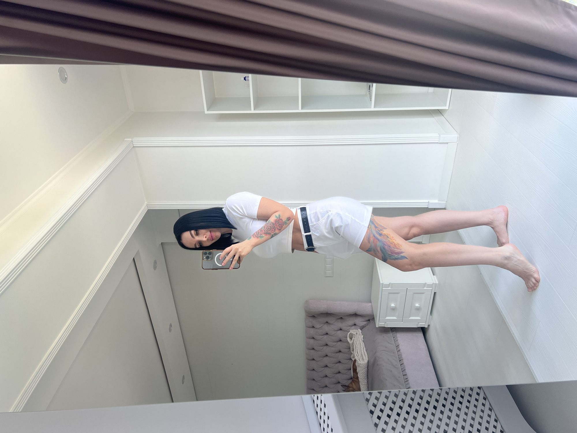 In an indoor setting, a young woman with black hair, wearing a white crop top and white shorts, stands by a full-length mirror, taking a selfie with her phone. Her left arm and left leg are adorned with colorful tattoos. The room appears to be a bedroom, reflected in the mirror, showing a tufted headboard bed with a blanket and a small dog lying on it, a white nightstand with two drawers, and a white shelving unit. A dark brown curtain is visible to the right of the frame.