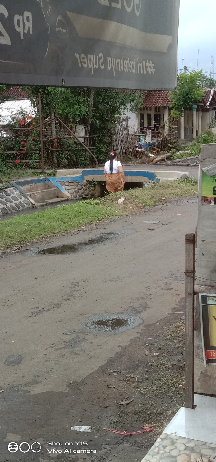 The scene depicts a rural or semi-urban setting in Maron Kulon, Indonesia, under an overcast sky with evidence of recent moisture on the ground. An unpaved road with numerous puddles occupies the foreground. A woman, seen from the back, stands on the left side of the road next to a small, low concrete bridge that spans a narrow water canal. She wears a white short-sleeved top and a light brown, patterned sarong-like garment, with her dark hair tied back. The canal features stone-clad retaining walls and concrete steps descending into the water on the left. The bridge has a distinct blue-painted edge along its concrete railing. Behind the canal, several single-story houses with red-tiled roofs are visible amidst dense green foliage. One house has white walls and dark-framed windows/doors. Scattered logs and debris are present near the structures. In the far background, utility poles and overhead lines are discernible against the sky. A banner with mirrored text, partially legible as "#iniketeknyasuper" and "Rp", hangs over the upper portion of the frame. The lower right foreground includes part of a wooden post and a small sign or advertisement. A white plastic object and red string lie on the ground in the immediate foreground.
