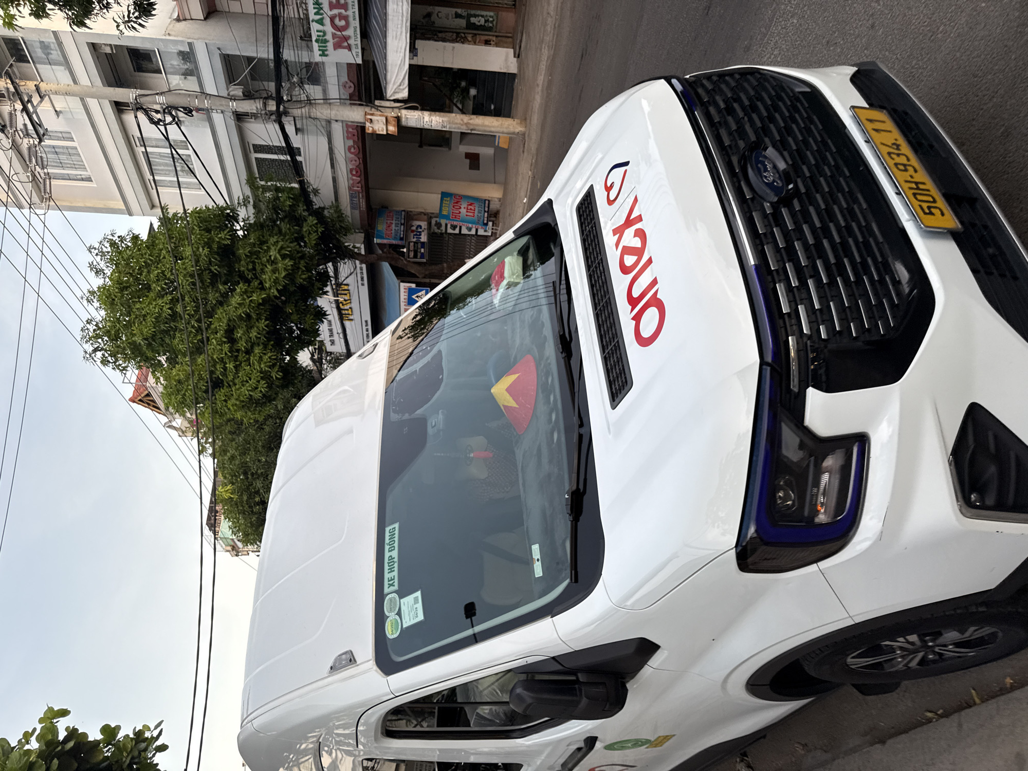 The image shows a white van parked on a street in Nha Trang, Vietnam, with a red and black "anex" logo on the hood and a yellow license plate reading "50H-934.11". The van has blue accents around the headlights and a black grille with the Ford logo. A sticker with Vietnamese text, "XE HỢP ĐỒNG", is visible on the windshield, along with other stickers. Behind the van, buildings and trees line the street. A person is visible inside the van through the windshield. The sky is overcast, suggesting it is either early morning or late afternoon. The street appears to be in a commercial or residential area, with signs for businesses such as "HIỆU ÁNH NGHÊ" and "HOTEL NGỌC HI".