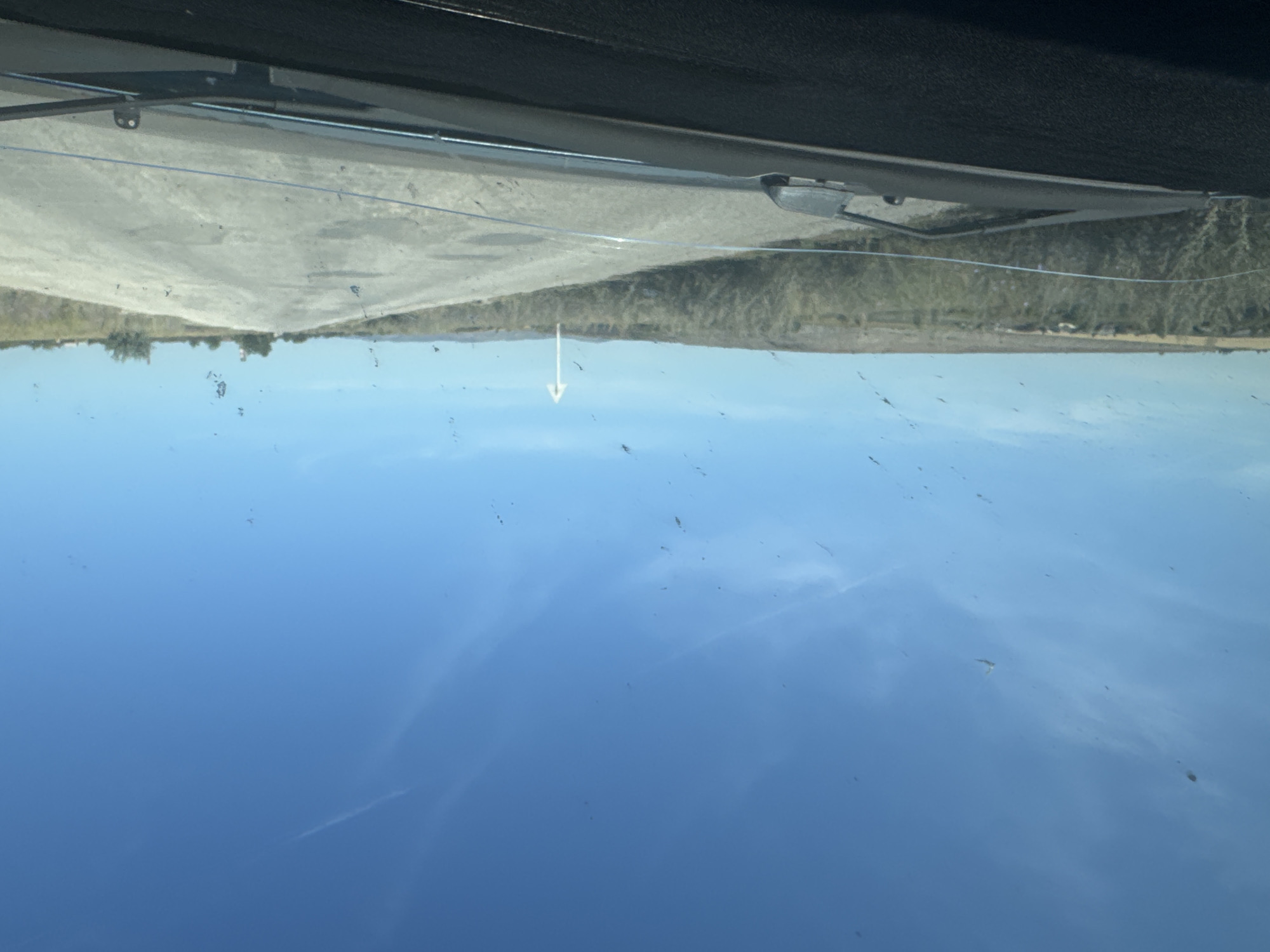 The image shows a view from inside a vehicle, looking out through a windshield. The windshield has numerous spots and streaks, suggesting it is dirty. A dark dashboard is visible in the foreground. The road ahead is paved but appears weathered and uneven, with some patches. The road is flanked by dry, scrubby vegetation and a flat, arid landscape under a clear blue sky. In the distance, there are some scattered trees and possibly buildings. A white directional sign with an arrow pointing to the right is visible along the roadside. The overall scene depicts a rural or semi-arid environment. No specific location in the city of Sibay, Russia is identifiable from the image.