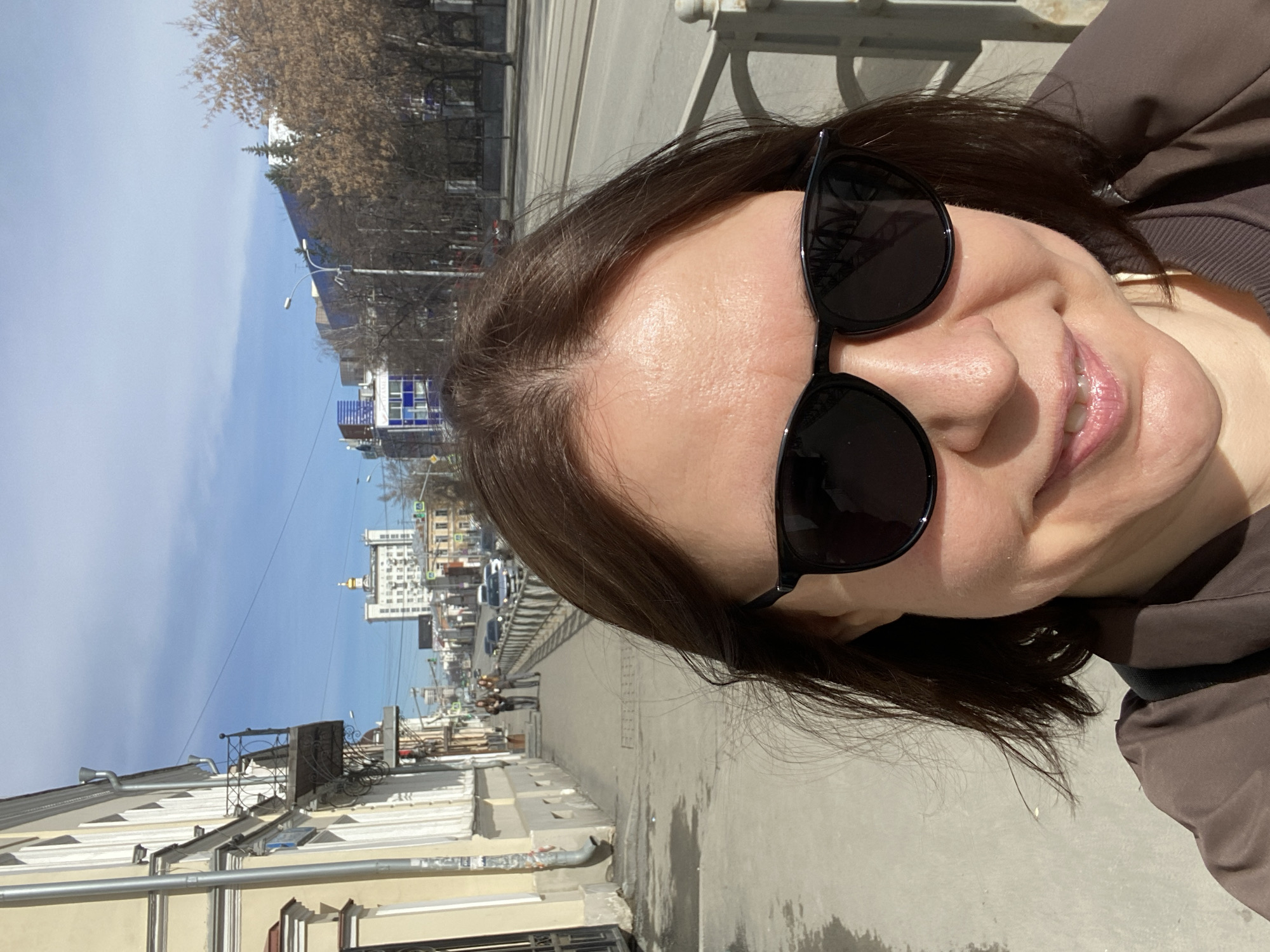 A woman with brown hair and sunglasses is taking a selfie on a city street. The street is lined with buildings and trees. In the background, there is a church with a golden dome. There are vehicles on the street. Based on the architectural style of the buildings and the presence of the church, the location appears to be Yekaterinburg, Russia.