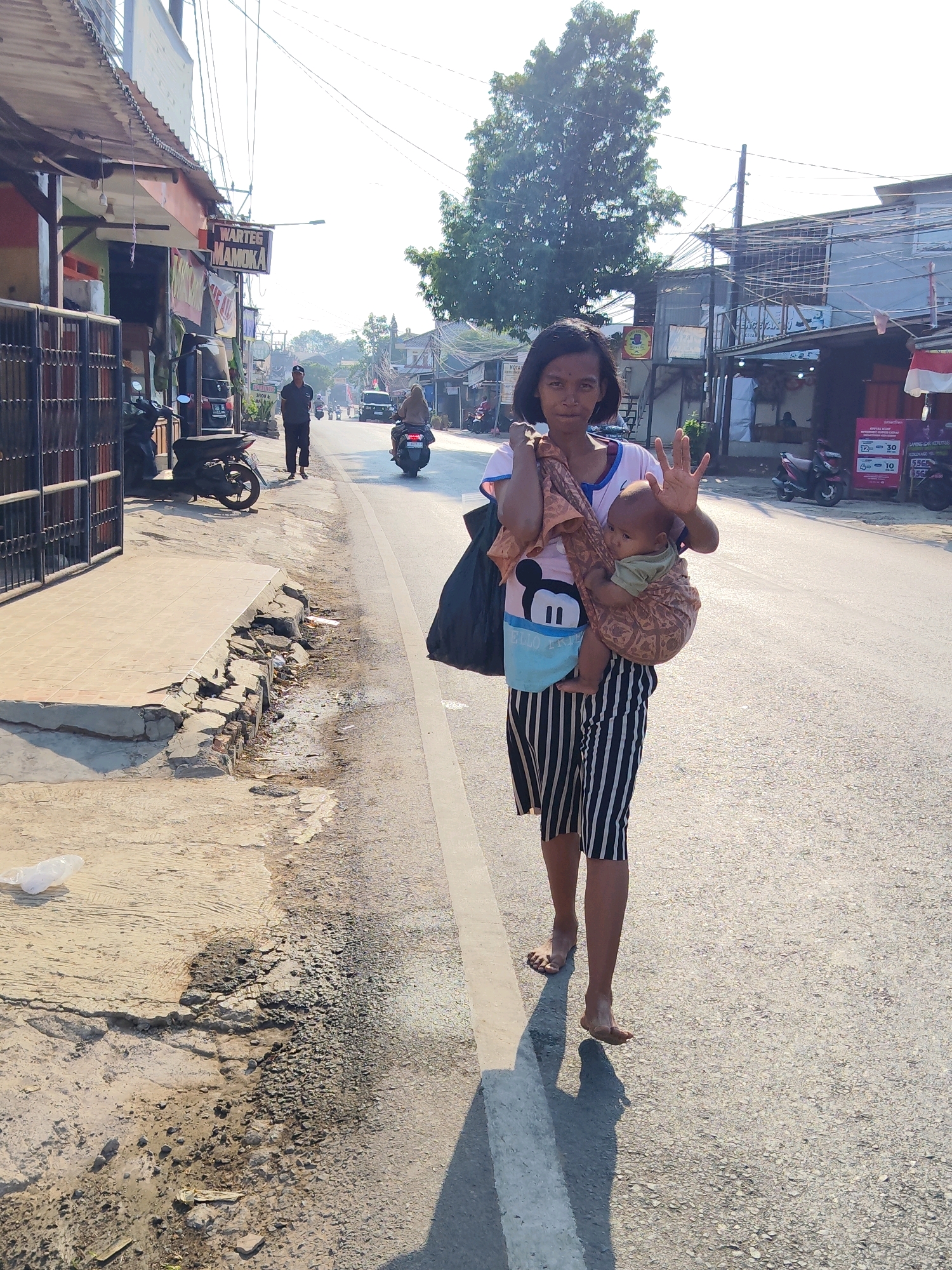 A young woman is walking barefoot on the side of a street in Purwakarta, Indonesia. She is carrying a baby in a sling on her chest. She is wearing a pink t-shirt with a Mickey Mouse graphic and black and white striped shorts. She is looking at the camera and holding up her hand, seemingly waving. A large black bag is slung over her left shoulder. The street is paved with asphalt, and there is a sidewalk on the left side of the image. The sidewalk appears to be damaged in some areas, with exposed bricks and debris. Several motorcycles and a few cars are visible on the road in the background, along with various buildings and shopfronts. A tall tree is situated behind the woman. The sun is shining, casting long shadows across the road and sidewalk.