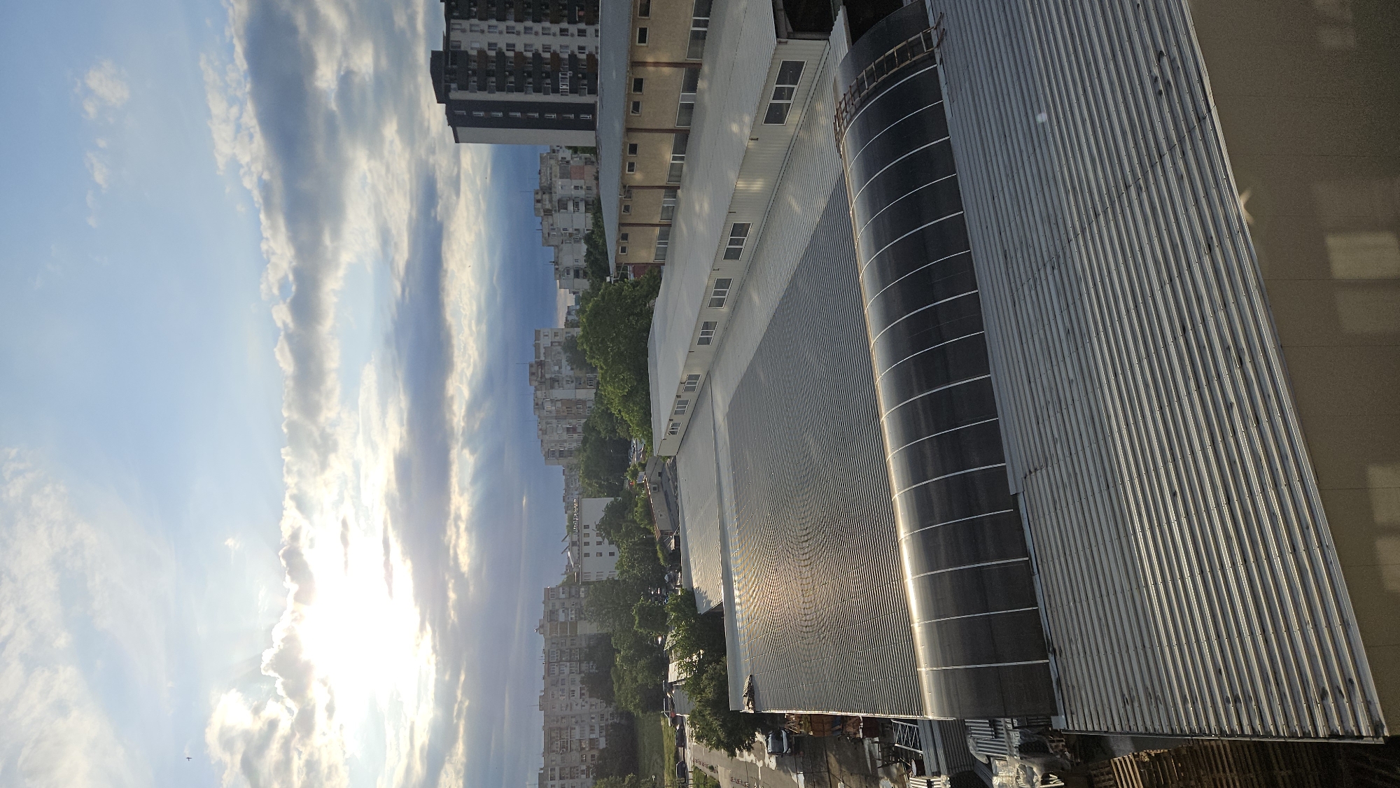 The image shows an aerial view of an urban landscape in Plovdiv, Bulgaria, taken during daylight under a sky with scattered clouds and bright sunlight. A large, modern building with corrugated metal siding and a curved, black cylindrical feature dominates the right side of the frame. To the left of this building are other structures, including residential buildings of varying heights and styles, some lined with trees. A street scene is visible below, featuring trees, parked vehicles, and a glimpse of a road. The photograph is taken from an elevated vantage point, suggesting a high floor of a building or a drone shot.