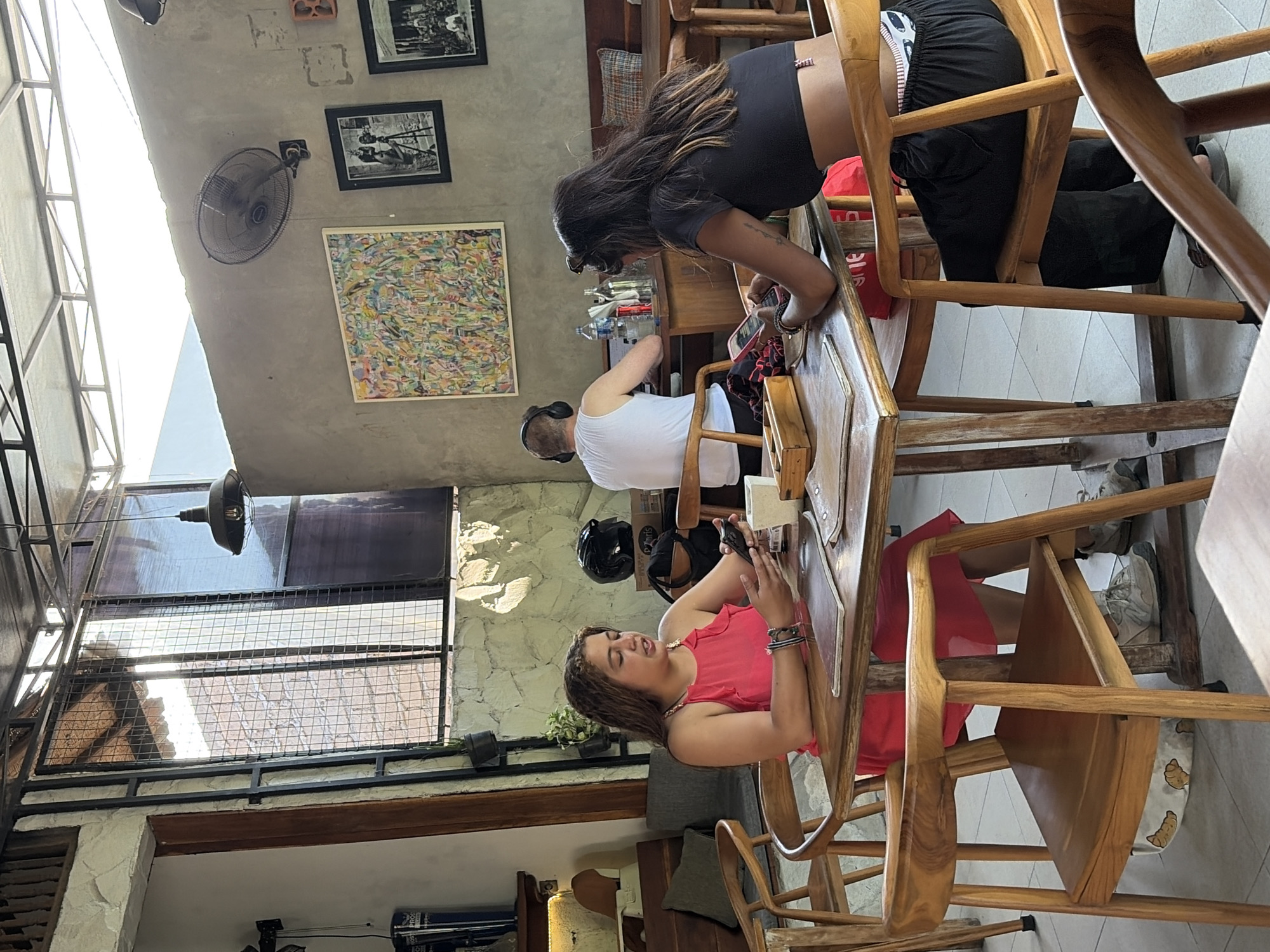 In a restaurant in Kubu, Indonesia, two women are seated at a wooden table. The woman on the left, wearing a pink sleeveless dress, is holding a mobile phone and appears to be speaking. The woman on the right, in a black crop top and black pants, is also looking at her phone. A man in a white tank top and headphones is visible in the background, seated at another table. The restaurant has an open-air design with a corrugated metal roof supported by a metal frame. The walls are made of textured concrete and stone. Artwork and framed photographs hang on the wall. Wooden chairs and tables are arranged throughout the space, and a large oscillating fan is mounted on the wall. Sunlight streams in through large windows, creating bright areas and shadows.