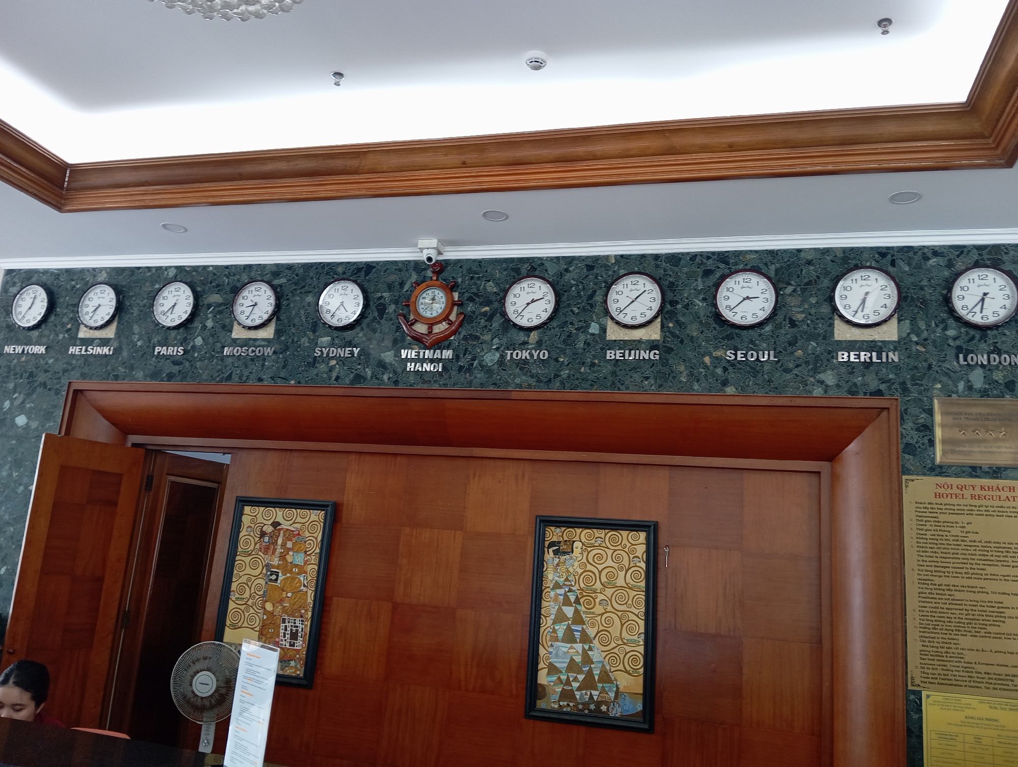 The image shows the reception area of a hotel, likely in Nha Trang, Vietnam. A line of world clocks is mounted on a dark green marble wall, displaying times for New York, Helsinki, Paris, Moscow, Sydney, Vietnam Hanoi, Tokyo, Beijing, Seoul, Berlin, and London. A security camera is visible above the "Vietnam Hanoi" clock. Below the clocks, a reception desk made of polished wood is partially visible, with a woman with dark hair sitting at the far left. A large, oscillating fan stands on the desk. Two framed artworks are hung on the wooden wall behind the desk. One artwork features abstract gold swirls and figures, while the other depicts a woman in a patterned dress. A printed hotel regulation document is affixed to the marble wall on the right. The ceiling features recessed lighting and a decorative chandelier or light fixture. The overall environment suggests a formal and well-maintained hotel reception.