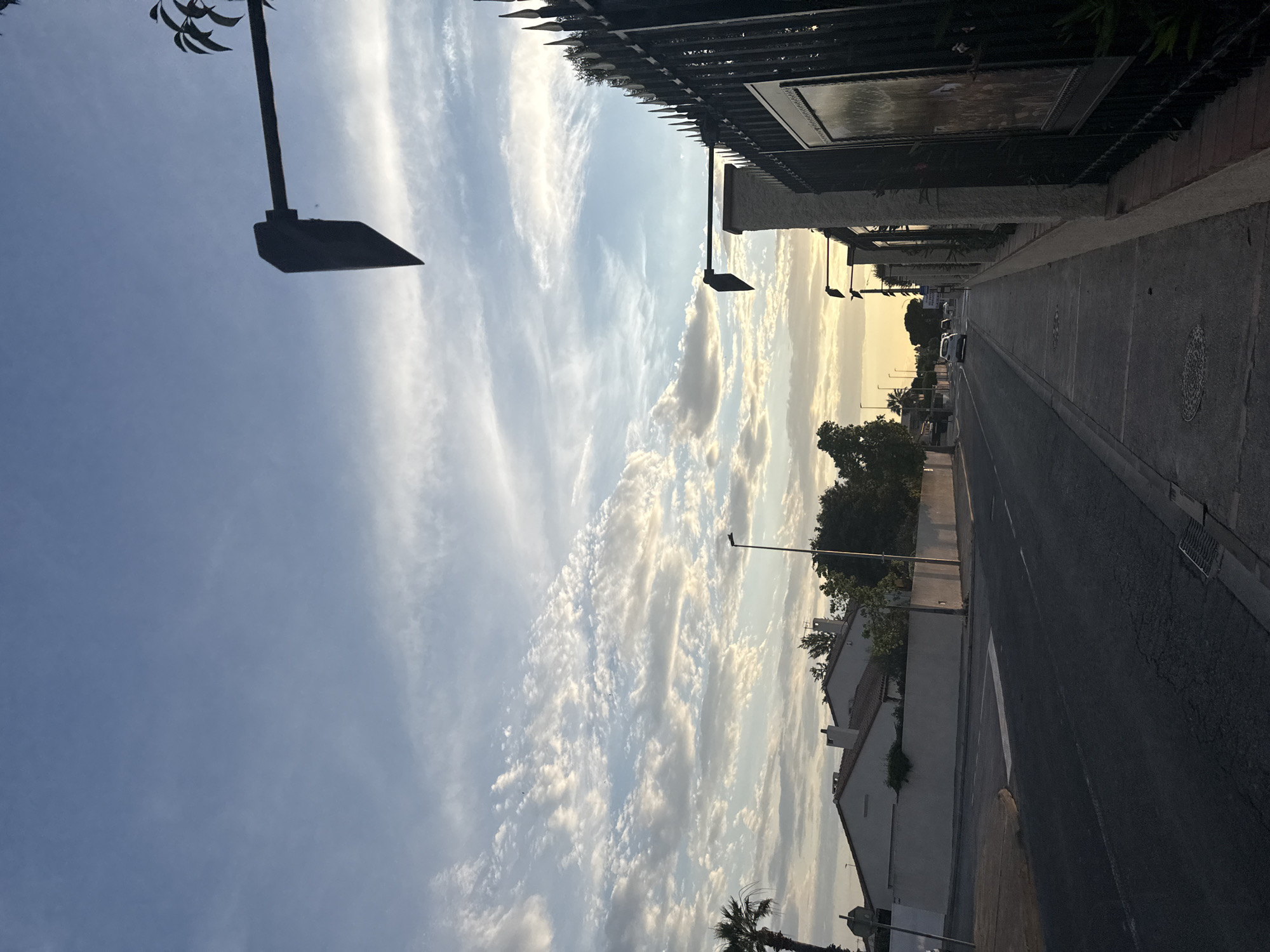 This is an eye-level, outdoor, medium shot of a street in Pia, France at dusk. The sky is a gradient of blue with scattered white clouds. On the left side of the road are houses and palm trees. The road is asphalt with white lane markings. A sidewalk runs along the right side of the road, bordered by a black metal fence and a stone wall. Two cars are visible on the road, one white and one dark colored. Traffic lights are visible suspended over the road. The overall lighting suggests it is late afternoon or early evening, with the sun setting.