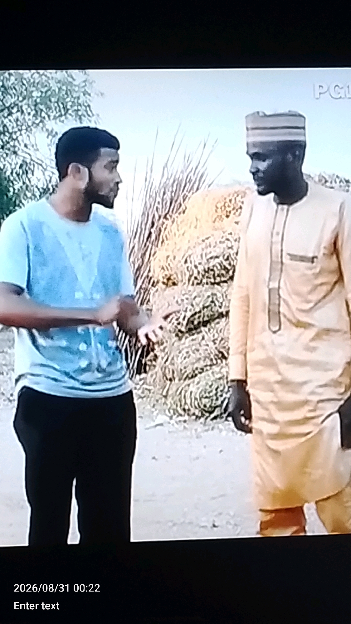 Two men are standing outdoors, possibly in Lokoja, Nigeria, with a blurry background of vegetation and a pile of what appears to be hay or dried grass. The man on the left wears a light blue t-shirt and dark pants. He has dark hair and a beard and is gesturing with his hands. The man on the right wears a light yellow traditional garment and a striped cap. He has dark skin and is looking towards the man on the left. The scene is captured at an angle, and the lower portion of the image displays a timestamp "2026/08/31 00:22" and the text "Enter text".