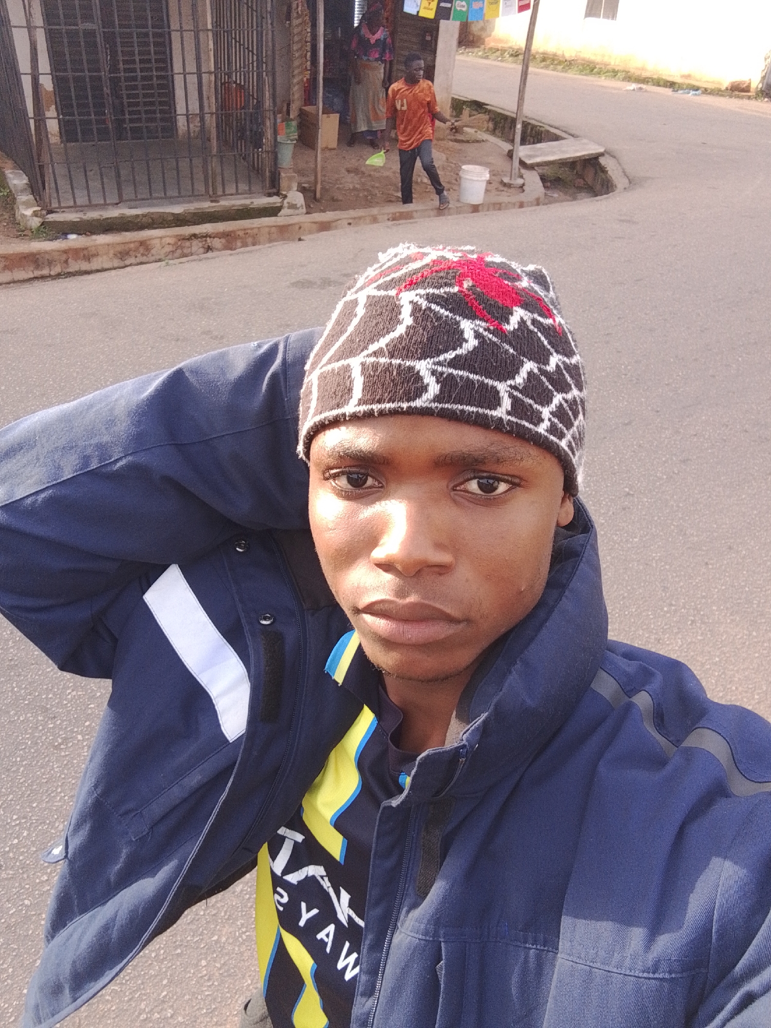 A young Black male is captured in a close-up, low-angle selfie taken outdoors on a paved street in Jos, Nigeria. He is wearing a dark blue jacket over a black, yellow, and blue patterned t-shirt. On his head, he sports a beanie with a red spiderweb design. His right arm is raised, with his hand resting on the back of his head. In the background, to the left, is a building with a metal grate. Further back, a young Black male in an orange t-shirt and jeans is walking. A white bucket is visible near him. The scene is likely a commercial or residential area.