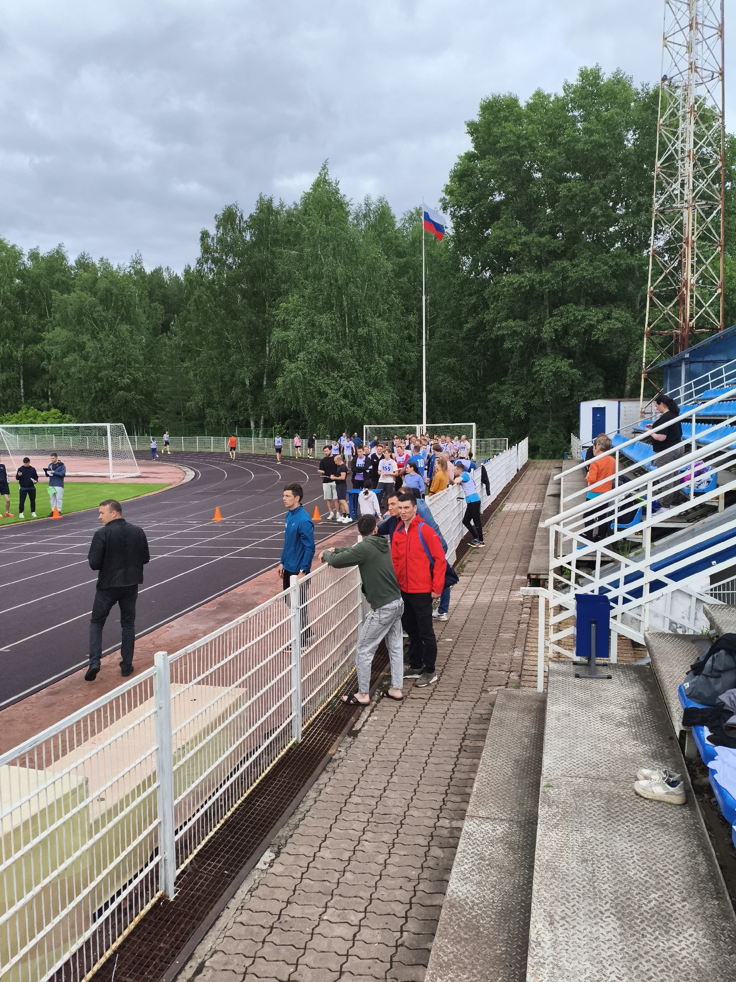 The image depicts a track and field stadium in Izhevsk, Russia, under a cloudy sky. A running track encircles a grassy field, with a soccer goal visible in the background. A crowd of people, likely participants and spectators, gathers along the sidelines and near the stands. Many of these individuals are dressed in athletic wear, suggesting they are participants in the event. A man in a black suit walks along the track, while others stand by the fence. The Russian flag flies from a pole near a metal tower. The stands are visible on the right side of the frame.
