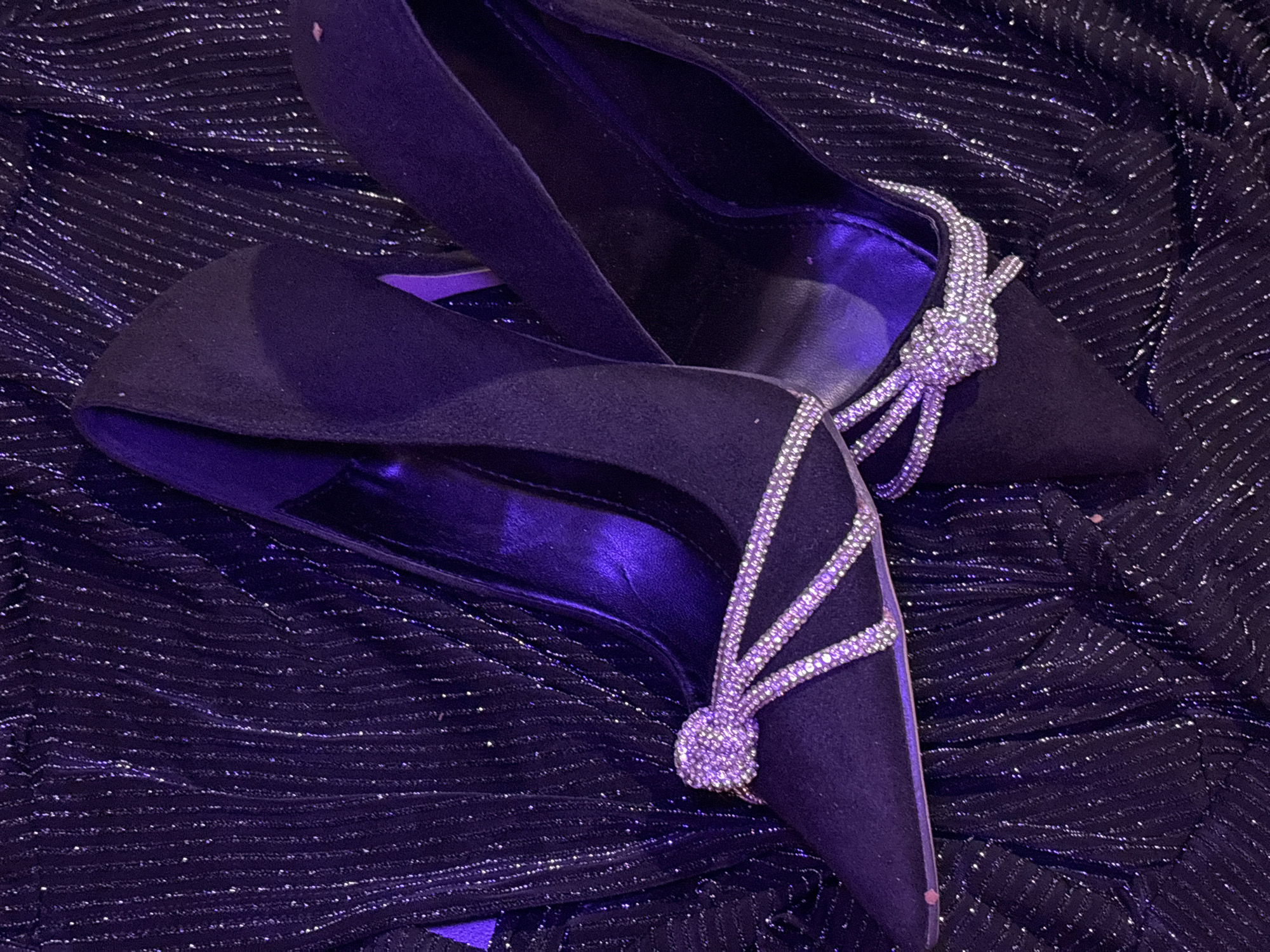 The image displays a close-up, top-down view of a pair of black high-heeled shoes, or pumps, resting on a dark, shimmering fabric.
The shoes are pointed-toe and appear to be made of a matte material, possibly suede or velvet, giving them a luxurious texture. Each shoe is adorned with an elaborate embellishment on the vamp, featuring a central knot intricately crafted from numerous small, sparkling rhinestones. From this knot, several rhinestone-studded straps fan out towards the sides of the shoe, adding a decorative and glamorous touch. The interior of the shoes is dark, with some faint traces of dust visible on the insoles in the deeper sections.
The background consists of a dark fabric, likely black or deep indigo, characterized by a textured, crinkled surface with numerous horizontal or slightly wavy lines of sparkling, metallic threads. These threads catch the light, creating a glittery effect that complements the rhinestones on the shoes.
The overall scene is dimly lit, with a prominent purple hue cast over the subjects and background, suggesting artificial lighting conditions that enhance the dramatic and opulent feel of the composition. No activity is taking place; the shoes are static and appear to be posed for display. There is no visible text in the image.