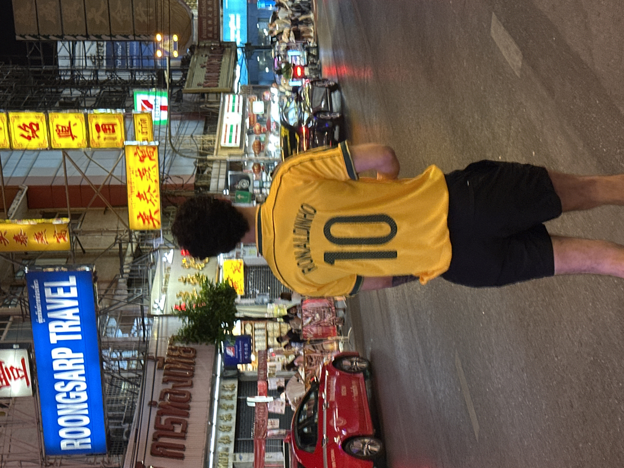 The image presents an overhead view of a person from behind, walking or standing on a lively city street at night in Bangkok, Thailand. The subject is wearing a yellow short-sleeved jersey with "RONALDINHO" and the number "10" emblazoned in black on the back, complemented by black shorts. Their dark, curly hair is visible.
The scene is an illuminated urban environment, characterized by a dense array of commercial buildings and a multitude of brightly lit signs in the background. The street itself appears to be asphalt, showing some wear and faint white road markings. Various vehicles, including a red car partially visible in the foreground and other cars and motorcycles with glowing taillights further down the street, suggest active traffic.
The most prominent feature of the setting is the abundance of illuminated signage. Many signs feature vibrant yellow backgrounds with red Chinese characters, stacked both horizontally and vertically. A clear blue rectangular sign displays "ROONGSARP TRAVEL" in white English text, with Thai script above it. A recognizable green, white, and red "7-ELEVEN" logo is also visible amidst the cluster of lights. The array of Chinese and Thai scripts on the signs, along with the architecture and bustling atmosphere, firmly places the location in an Asian city.
The time of day is clearly night, indicated by the darkness of the sky and the glowing illumination from the numerous commercial signs and streetlights. The overall impression is that of a vibrant, energetic commercial district after dark, with the person seemingly navigating or observing the urban nightlife.