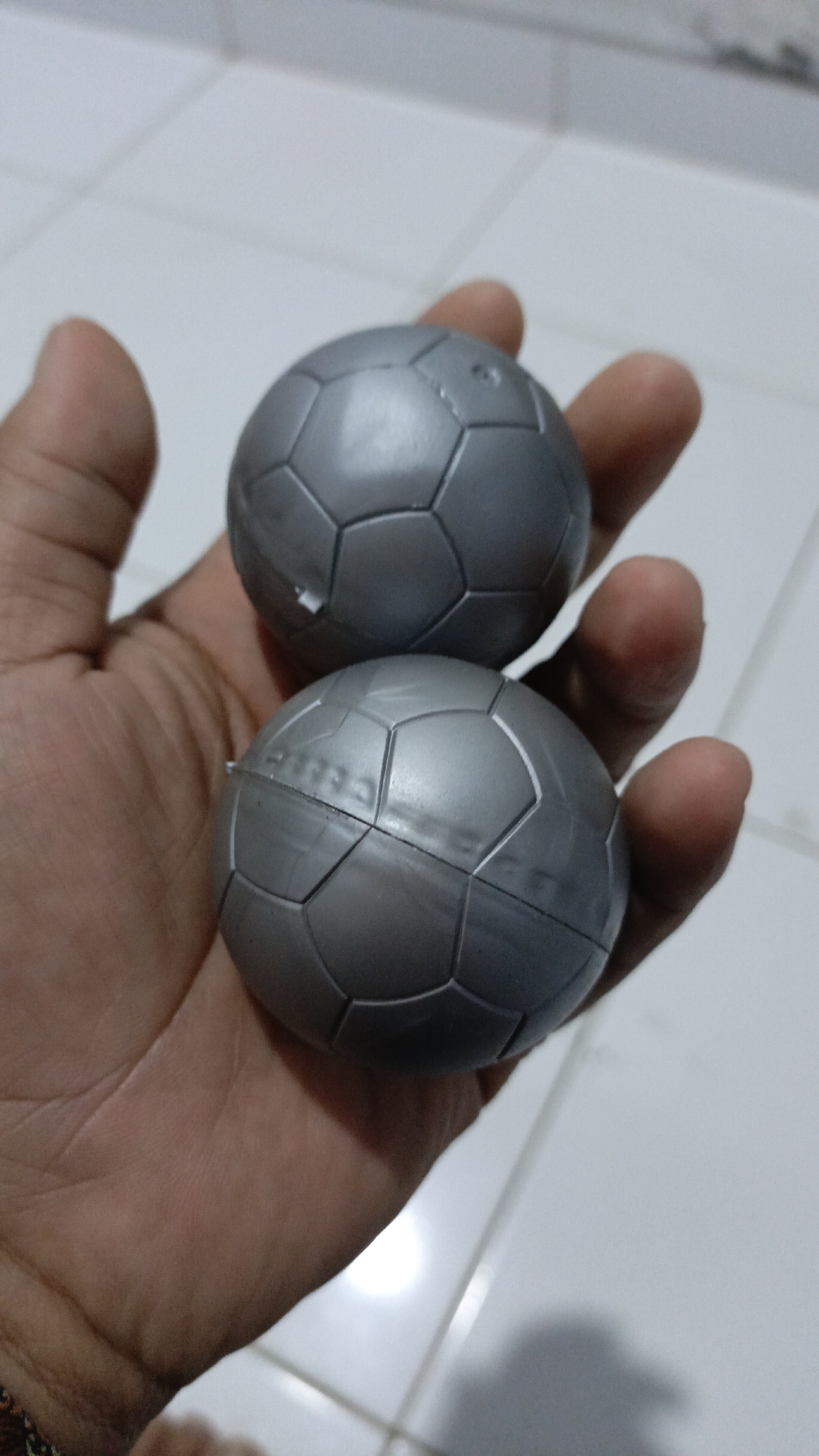 Two small, silver, toy soccer balls are held in a person's left hand. The balls are hexagonal and pentagonal panel patterned. The background is a tiled floor. The image was taken on March 26, 2023, at 11:30 AM local time in Trihanggo, Indonesia.