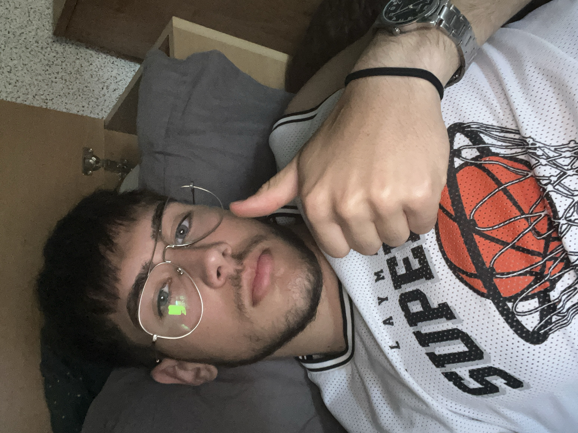 A young adult male is lying down, possibly on a bed or resting place, with his head resting on a gray pillow. He has short dark hair, a beard, and is wearing gold-rimmed glasses. He is wearing a white basketball jersey with black lettering and a basketball graphic. He is wearing a silver watch and a black elastic wristband on his left wrist. His left hand is raised with his thumb extended. The background shows parts of furniture, possibly a dresser or cabinet. The location is not discernible from the image.