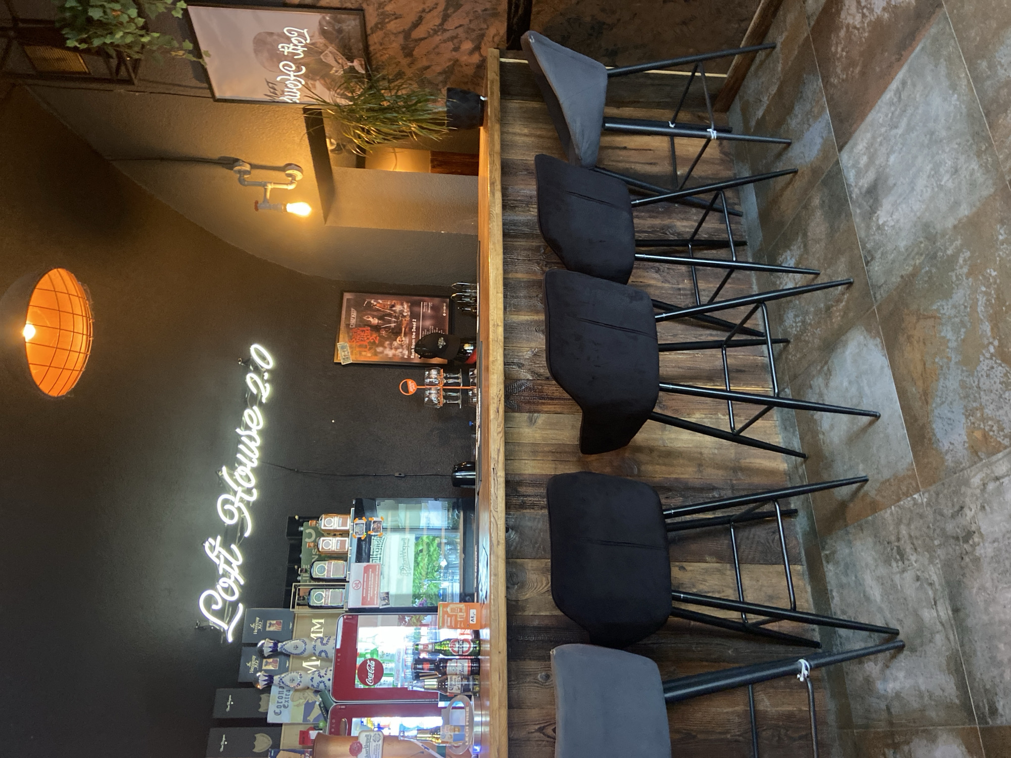 The image depicts the interior of a bar or restaurant, specifically the bar area, located in Bolesławiec, Poland. A neon sign on the dark wall above the bar reads "Loft House 2.0". The bar itself is made of wood, with a counter top at approximately waist height. Mounted on the wall behind the bar are various bottles, including what appear to be spirits and possibly some beer. A red Coca-Cola branded refrigerator is also visible, containing beverages. In front of the bar, facing it, are four high stools. Two are dark grey, and two are black. The floor is tiled with a pattern resembling aged concrete or stone. To the right of the bar, a large potted plant stands near a framed picture on the wall, which seems to be a poster with text. The lighting appears dim, with an orange-toned light fixture hanging from the ceiling and a smaller exposed bulb fixture on the wall. There are no people visible in the image.