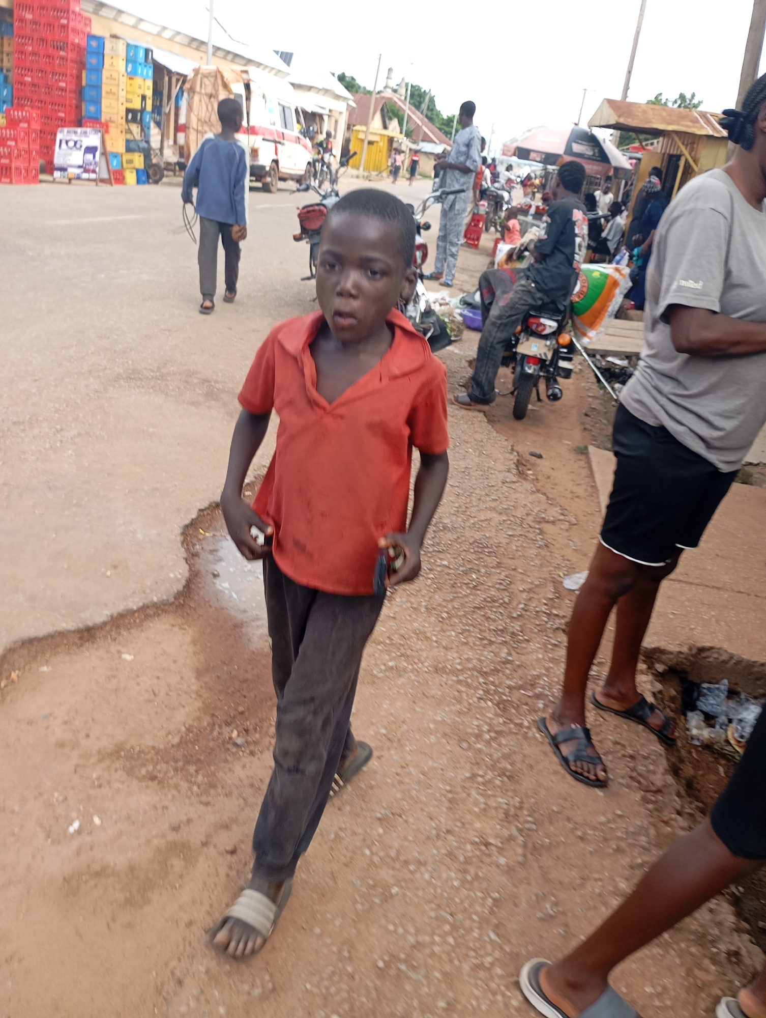 The image captures a street scene in Shendam, Nigeria. In the foreground, a young boy wearing an orange t-shirt and dark pants walks towards the camera. He is barefoot. To his right, another person, partially visible and wearing black shorts and a grey t-shirt, stands with their feet in sandals. In the mid-ground, several people are gathered around motorcycles and stalls. A person in a blue shirt and grey pants walks away from the camera, carrying a coiled wire and what appears to be a small object. Behind them, a white and red vehicle is parked. The background shows a street lined with various stalls and buildings, with stacks of colorful crates, likely containing beverages, visible on the left. The ground is unpaved and dusty.