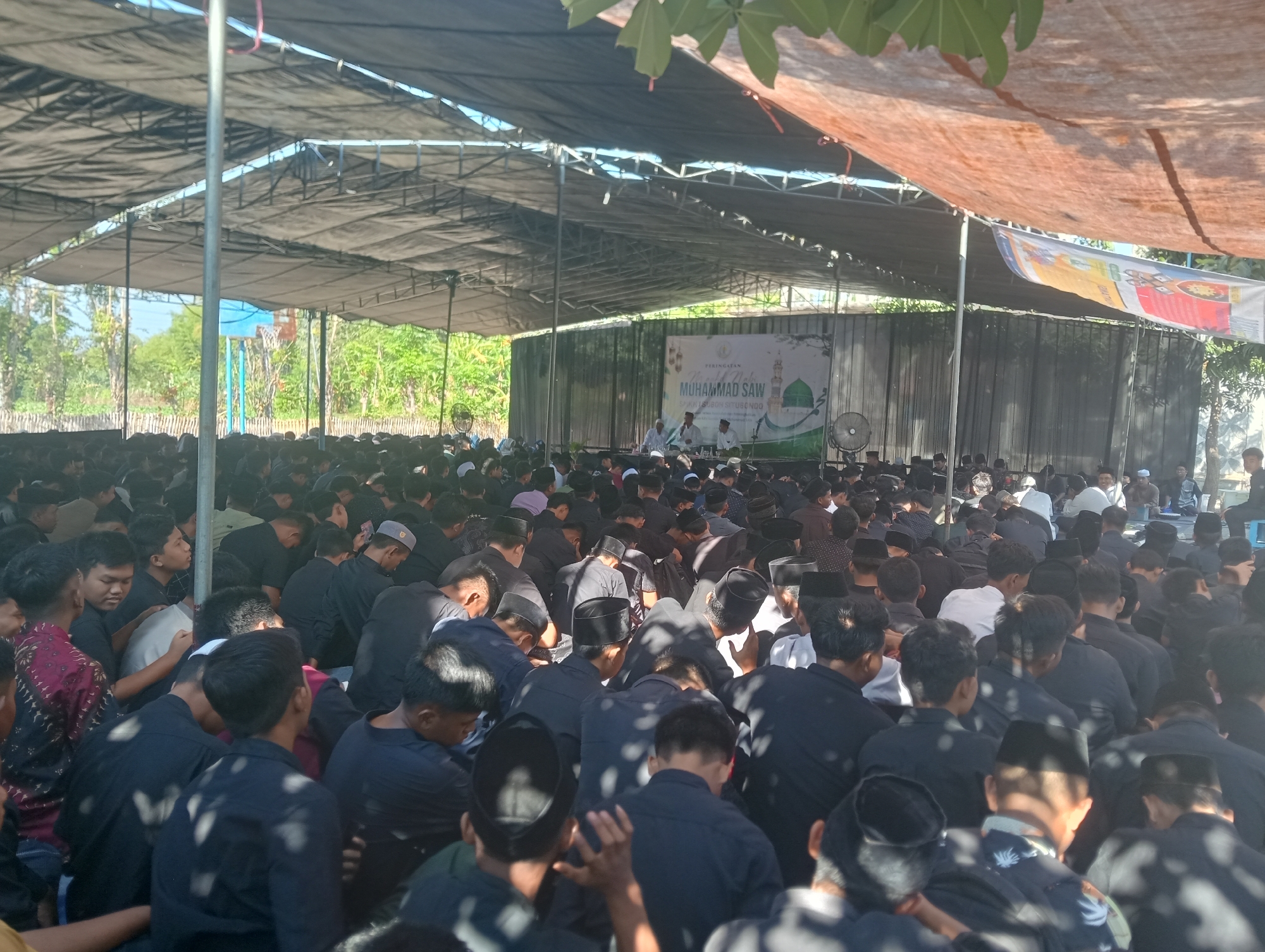 The image depicts an outdoor gathering of a large group of people, predominantly young men, seated on the ground under a makeshift canopy. They are attending an event in Buduan, Indonesia, indicated by a banner in the background that reads "PERINGATAN MAULID NABI MUHAMMAD SAW" and "BUDUAN SITUBONDO". The crowd is mostly dressed in black attire, with many wearing peci or kopiah, traditional caps.
The focus of the event appears to be a stage or platform at the back of the gathered crowd, where several men are seated, some speaking or engaged in conversation. A large banner is displayed behind them, featuring an illustration of a mosque and Islamic calligraphy. Two electric fans are visible on either side of the stage, suggesting a warm climate.
The environment is characterized by the natural elements surrounding the venue. Trees and greenery are visible beyond the canopy, and the sky is a clear blue. The overall atmosphere suggests a community event or religious observance. The lighting indicates it is daytime, with sunlight filtering through the gaps in the canopy, creating dappled shadows on the crowd. The people are mostly facing towards the stage, indicating their engagement with the proceedings.