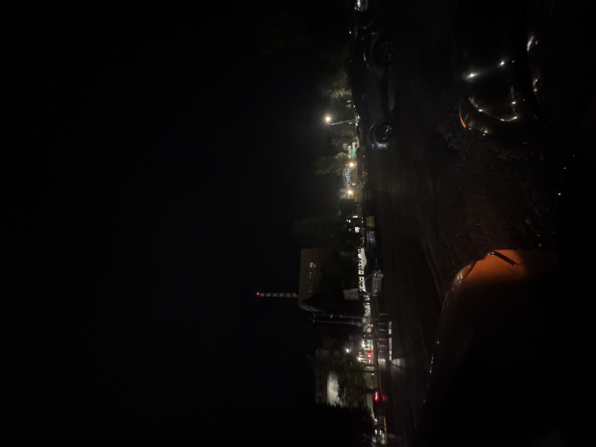 The image depicts a nighttime scene in Stara Zagora, Bulgaria. A street lined with parked cars and buildings stretches into the distance, illuminated by streetlights. A tall structure with red and white stripes is visible. A vehicle is in the foreground.