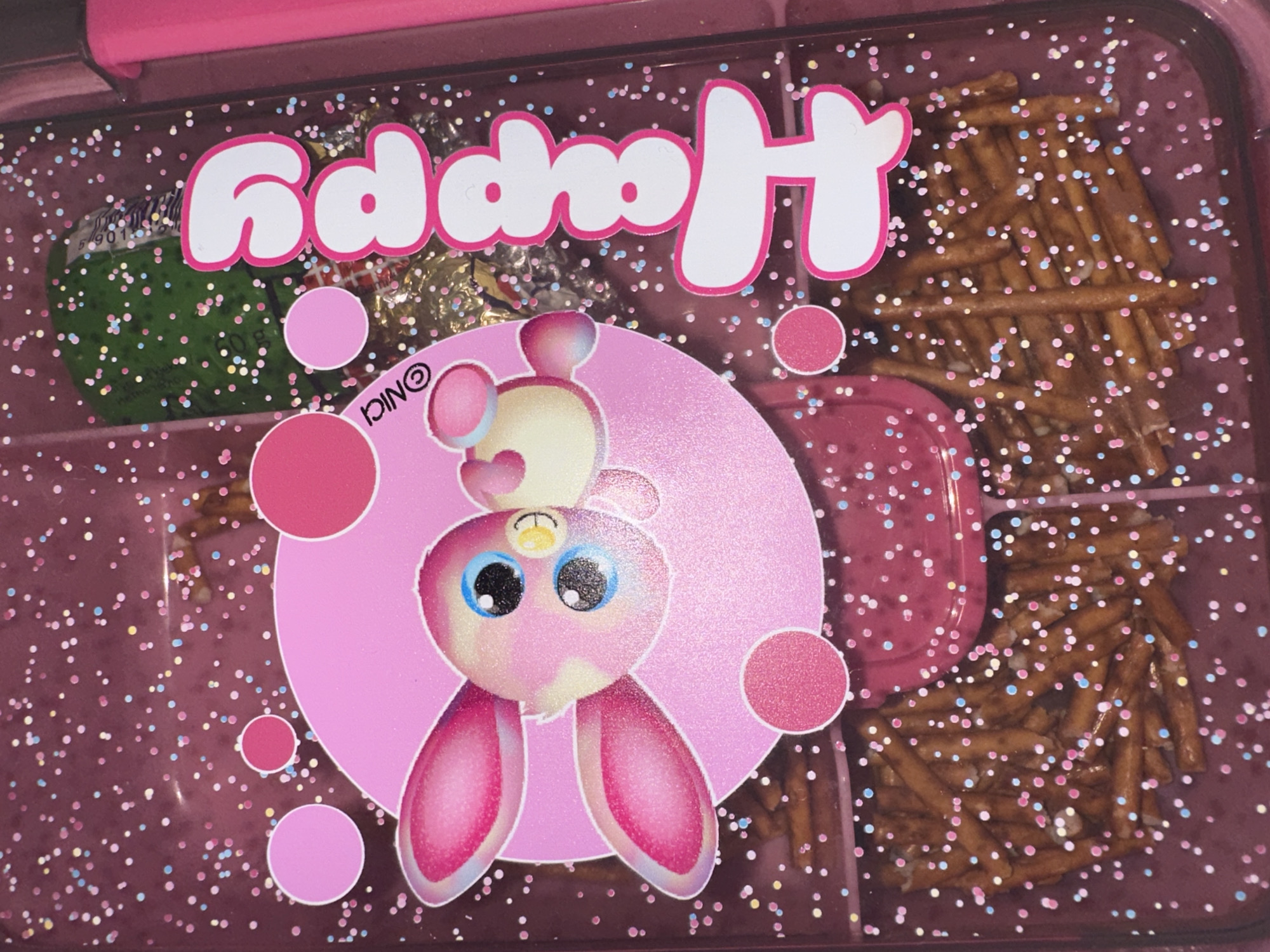 The image shows a pink lunchbox filled with snacks. The lunchbox has a clear lid with a pink border and is decorated with the word "Happy" in large white letters with pink outlines. There is also a pink round sticker featuring a cartoon bunny with big blue eyes and floppy ears. The lunchbox contains various snacks, including a wrapped green candy, foil-wrapped chocolates, pretzels, and a small pink container. The background is speckled with colorful dots, adding to the playful and childlike aesthetic. The location in the city is Unknown, Unknown.