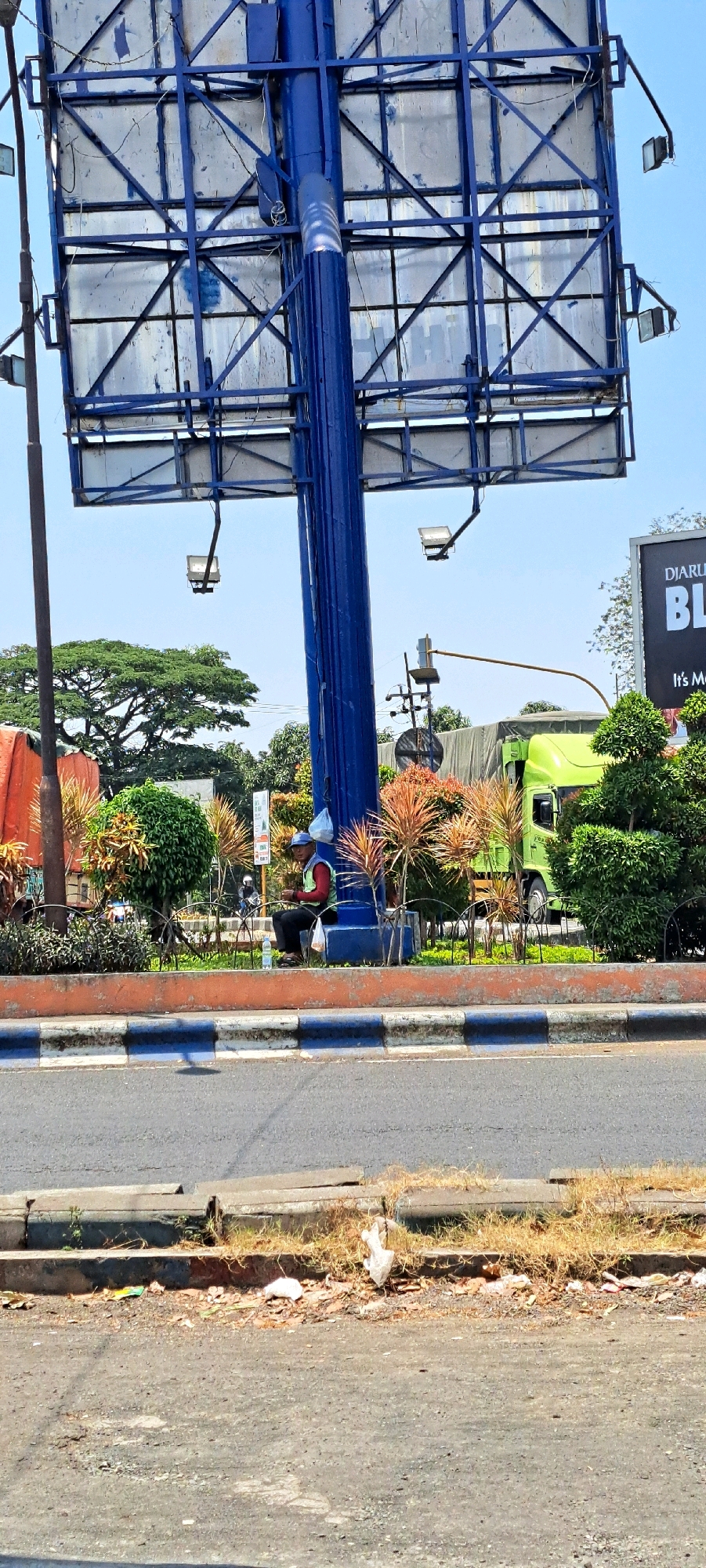 In Pandaan, Indonesia, a man wearing a blue cap, a yellow vest over a red shirt, and dark pants sits on a blue structure near the base of a large, blue metal billboard. He is holding something in his hands. A white plastic bottle is on the ground next to him. The billboard is supported by a thick, blue metal pole. Various plants, including green shrubs and reddish-brown spiky plants, surround the base of the billboard. In the background, a bright green truck with a covered trailer is visible. A street with a concrete curb and dry grass lines the foreground. The sky is clear and blue.