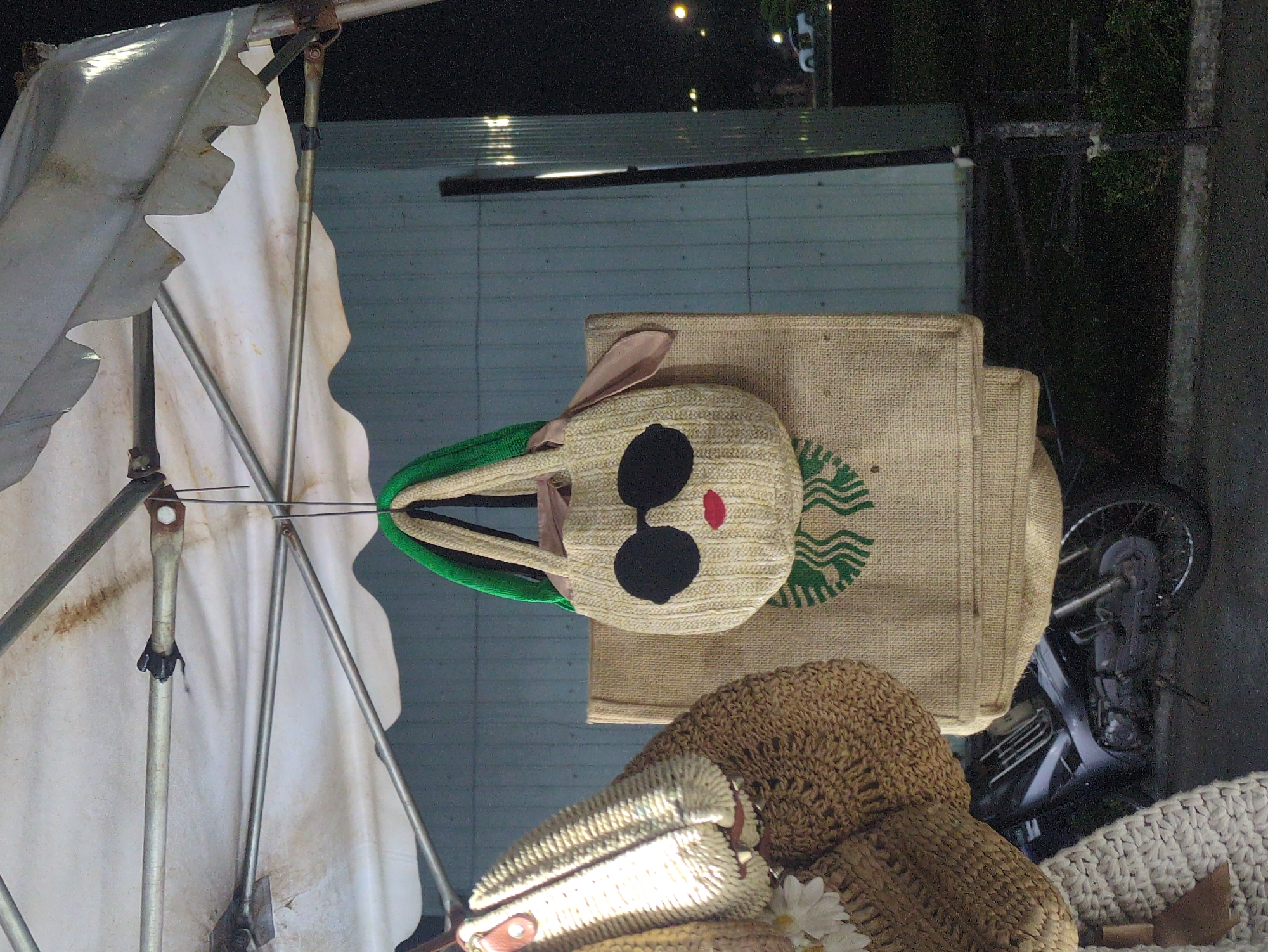 A roadside display in Quy Nhon Ward, Vietnam, features an open white umbrella with a tattered cover. Adjacent to the umbrella, a burlap bag with a green Starbucks logo sits atop straw hats. A motorcycle is partially visible in the background.