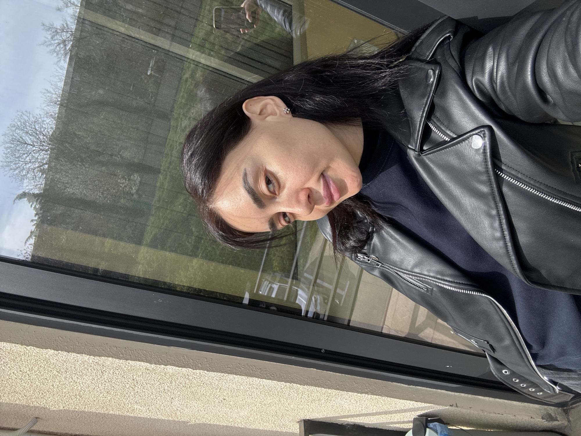 A woman stands indoors in Tbilisi, Georgia, near a window. She is wearing a black leather jacket and a dark top. She is holding a phone, visible in the reflection on the window. The window looks out onto trees and greenery. The exterior wall near the window has a textured surface.