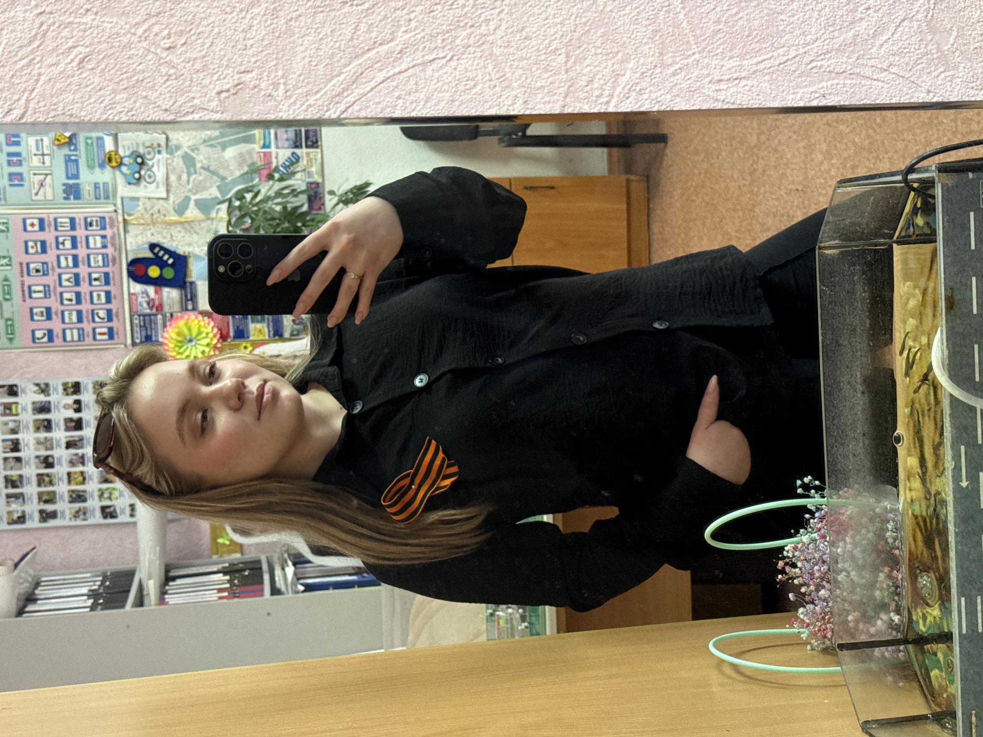 A young woman is taking a selfie in a mirror. She has light blonde hair and is wearing a black button-down shirt with a black and orange ribbon pinned to it. The background visible in the mirror includes a whiteboard with maps and charts, a plant, and a wooden cabinet. There is also a desk visible with office supplies and a flower arrangement in a glass container. The photo appears to be taken indoors, possibly in an office or classroom. While it is not possible to ascertain the specific location in Chaikovsky, Russia from the image, these details suggest a workplace or educational setting.