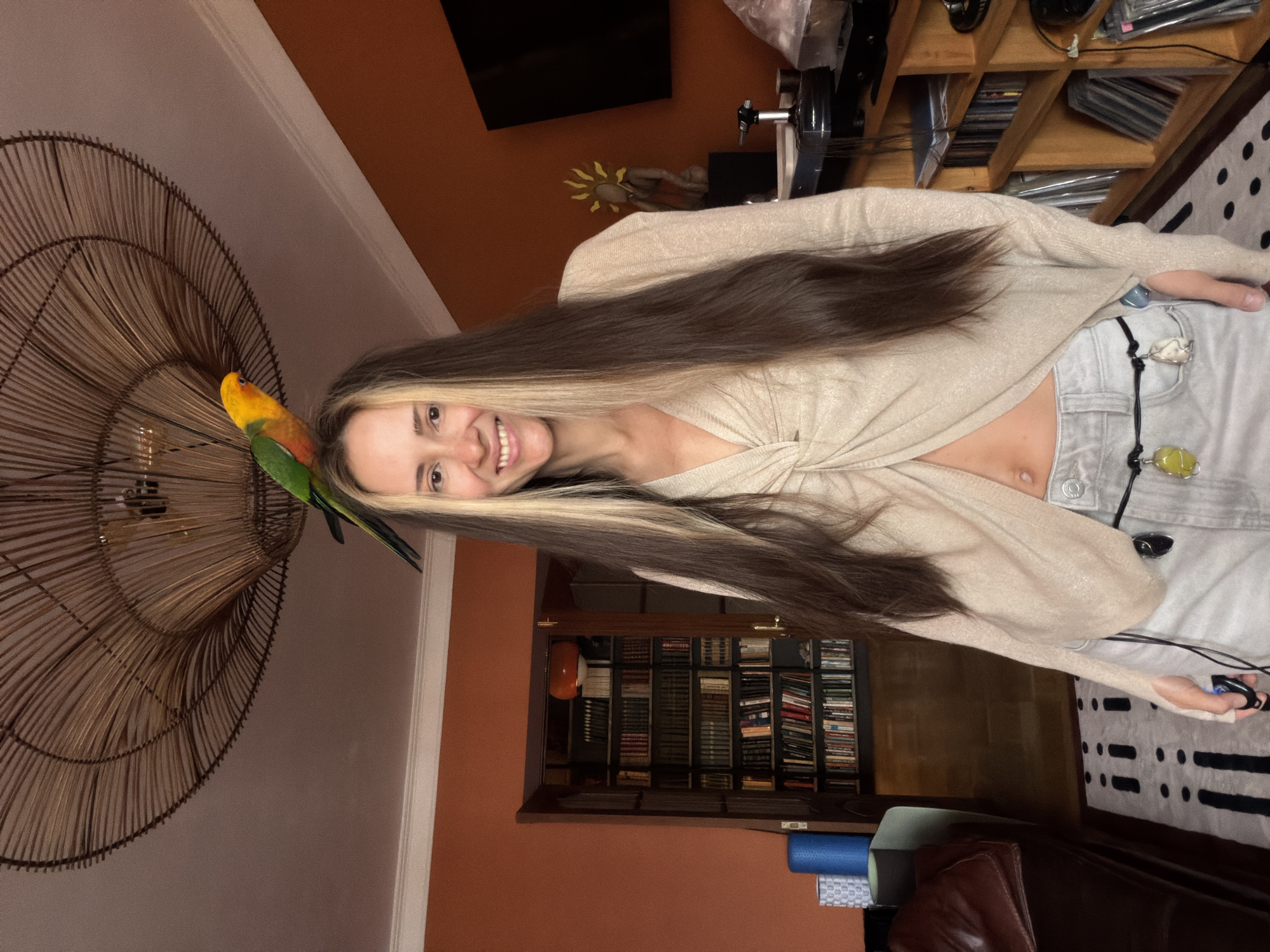 A woman with long, dark brown hair with blonde highlights stands in what appears to be a living room, with a parrot perched on her head. The parrot is colorful, with green, yellow, and orange feathers. The woman is smiling and wearing a light-colored, open-fronted top that reveals her midriff and light-wash jeans. She is accessorized with a belt adorned with several hanging pendants, one of which appears to be a yellow stone. In the background, a large, intricate woven light fixture hangs from the ceiling. To the left, a wall is painted orange and features a doorway leading to a room filled with bookshelves. To the right, a television is mounted on the wall, and a wooden shelving unit is filled with books and what appear to be CDs or records. The floor is covered with a rug featuring a black and white patterned design. The presence of a parrot and home decor suggest this is a domestic setting. The image does not contain any explicit or implicit indicators of the city of Khimki, Russia, or any other specific geographical location.