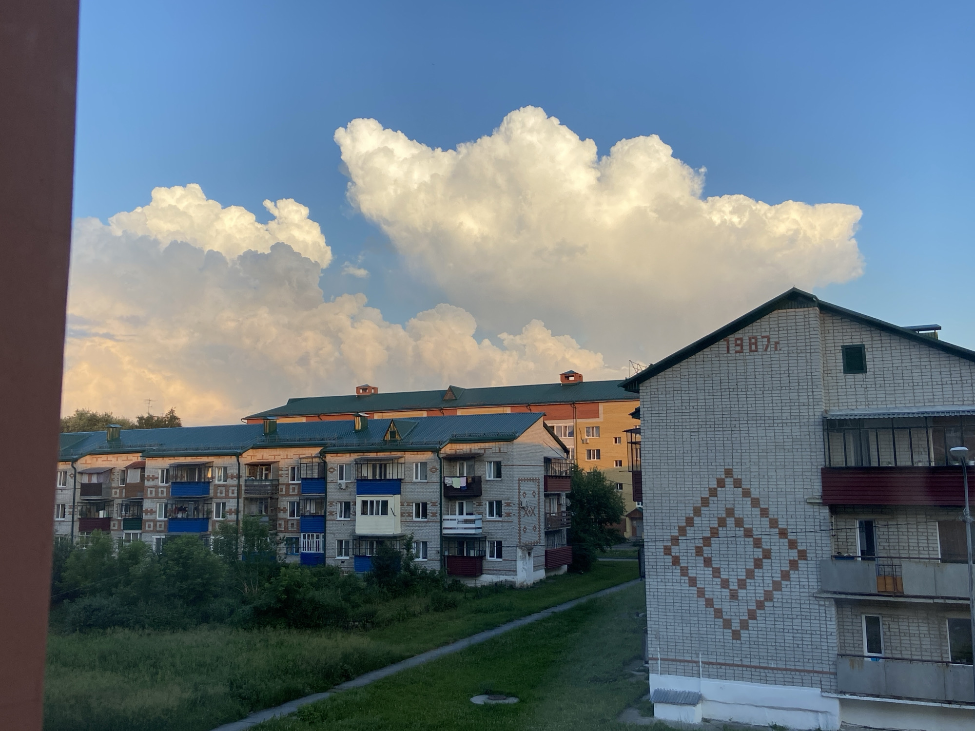 The image captures a view of apartment buildings in Zavodoukovsk, Russia, under a dynamic sky. The foreground is dominated by a large, fluffy cumulus cloud formation set against a bright blue sky, suggesting fair weather with potentially changing conditions. The sunlight illuminates the clouds, giving them a warm, golden hue.
Several apartment buildings are visible. The closest one on the right is a multi-story brick building with a distinctive diamond-shaped pattern made of darker bricks on its facade. The year "1987r" is also visible in red brick lettering on this building. Balconies with railings and windows are present on its side. Further in the background, a row of apartment buildings with green roofs and colorful balconies runs horizontally across the frame. These buildings appear older, with a utilitarian architectural style common in Soviet-era housing. Trees and green grass surround the buildings, indicating a residential area with some landscaping.
The time of day appears to be late afternoon or early evening, given the warm lighting and the long shadows that might be implied by the angle of the sun. The overall atmosphere is one of a typical day in a residential neighborhood, with a focus on the contrast between the imposing cloud formations and the everyday structures of urban life. There are no people or explicit activities depicted, but the scene suggests a quiet, lived-in environment. The visible text "1987r" on the building indicates its construction year.
