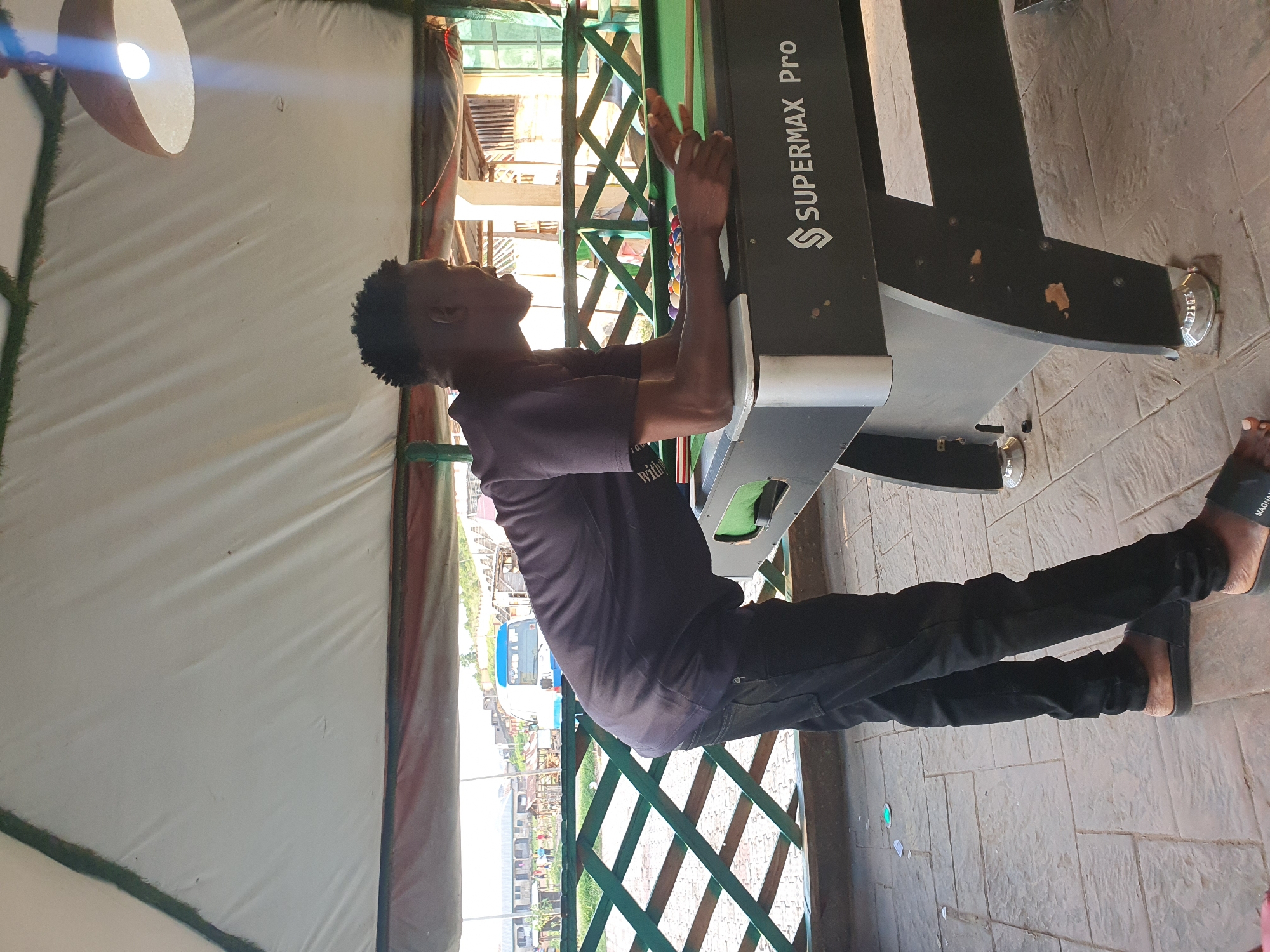 The media file shows a young man in Lokoja, Nigeria, in what appears to be an outdoor setting. He is leaning on a pool table, with his arms resting on the edge. He is wearing a dark t-shirt and black pants, and appears to be looking up and to the right. The pool table is dark green with the text "SUPERMAX Pro" visible on its side. The background shows a glimpse of a blue and white bus, and buildings. The lighting suggests it is daytime. The overall impression is of a casual, relaxed moment, possibly during a break or before a game.