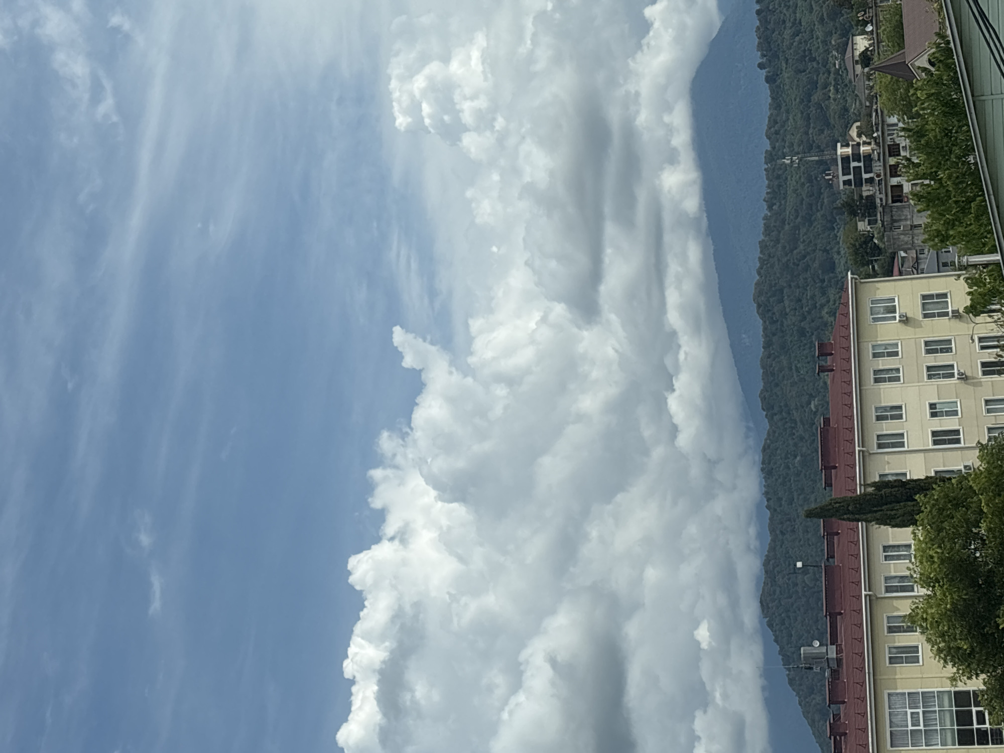 This is an eye-level, outdoor, medium shot of a cloudy sky over a town nestled in front of a mountain range. The sky is a light blue with wispy white clouds scattered throughout, and larger, cumulonimbus clouds dominating the middle ground. The mountains in the background are covered in dense green forest, with some mist obscuring the peaks. In the foreground, a light-yellow, multi-story building with a red roof is visible. It has numerous windows and what appear to be air conditioning units. Trees and other buildings are also present in the lower part of the frame. The scene is calm and depicts a typical day in a town near mountains. The location is identified as Vesyoloye, Russia.