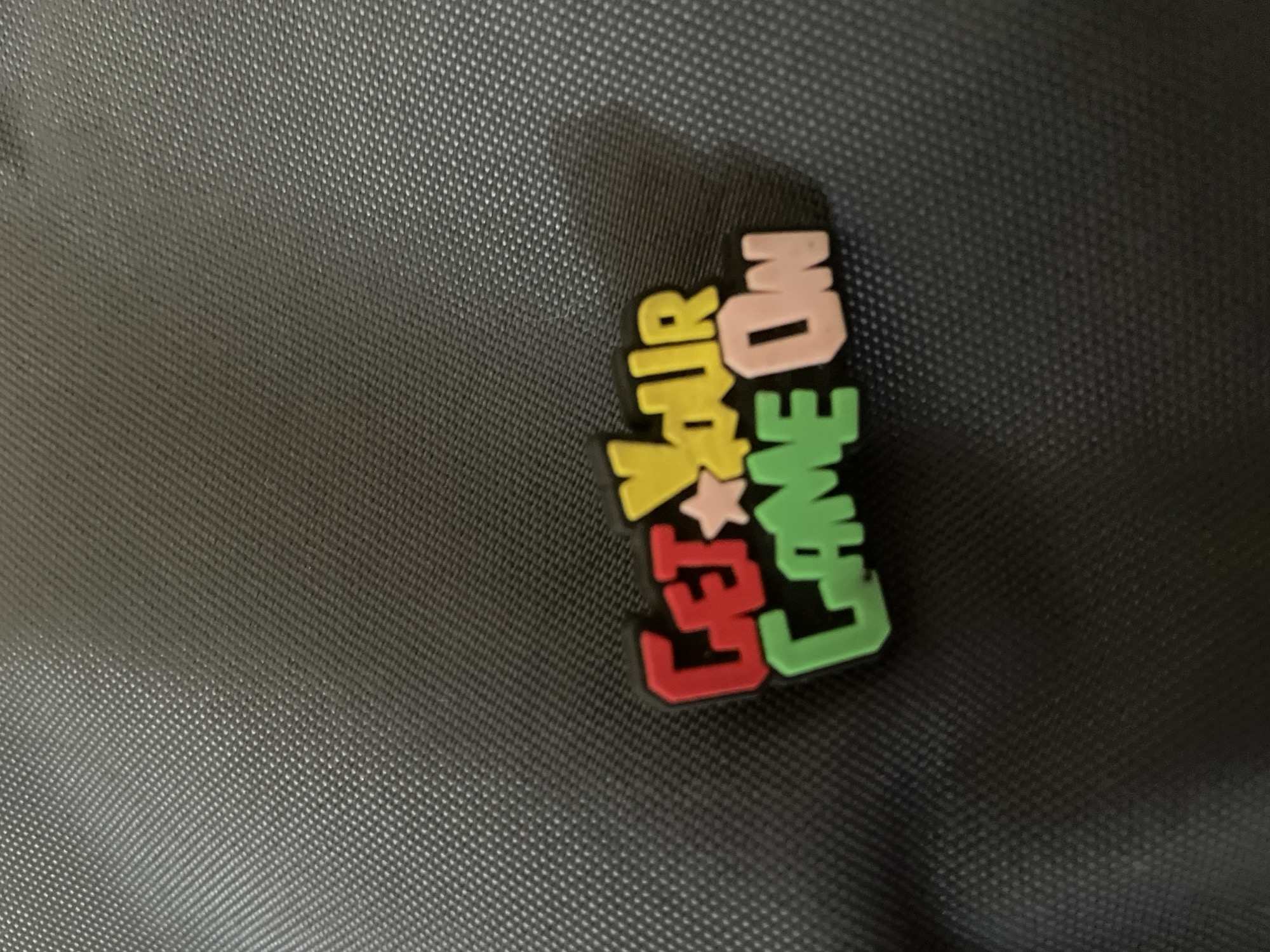 A colorful, multi-lettered charm is the central subject of this close-up, slightly angled photograph. The charm, designed with raised, block-style letters, spells out the phrase "LET YOUR GAME ON." The word "LET" is rendered in red, "YOUR" is in yellow with a small, light-pinkish star replacing the dot above the 'I', and "GAME ON" is divided, with "GAME" in vibrant green and "ON" in light pink. The letters appear to be made of a flexible material like rubber or silicone.
The charm rests on a dark, textured surface, possibly a synthetic fabric or molded plastic. This background has a distinct, repeating pattern of small, raised dots or squares, giving it a structured, dimpled appearance. The lighting is relatively soft and even, highlighting the texture of both the charm and the background.
There are no people or specific activities taking place; the image is a static portrayal of the object. No explicit location cues are visible within the image itself to suggest "Baranovka, Russia," which is provided as external context. The orientation of the charm is diagonally across the frame, with "LET" positioned towards the bottom left and "ON" towards the upper right. The overall impression is a vibrant, motivational phrase presented on a utilitarian, dark backdrop.