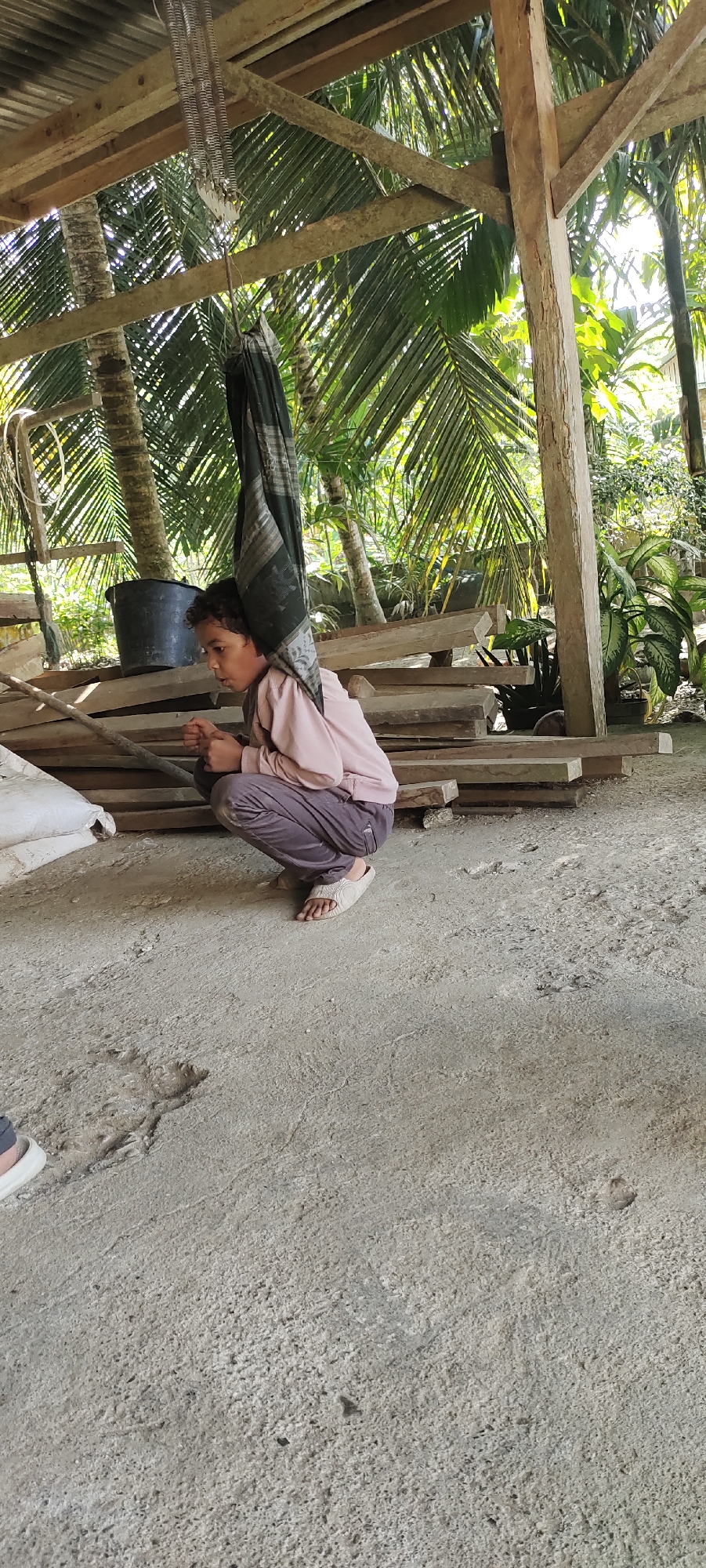 The image shows a person squatting on a concrete floor under a wooden structure. The person is wearing a pink long-sleeved shirt and gray pants with light-colored sandals. A dark-colored cloth is draped over their head and shoulders, hanging from the structure above. Behind the person, there are stacks of lumber and a large black bucket. In the background, lush green trees and plants are visible. The location is under a wooden structure in what appears to be a yard or outdoor area.