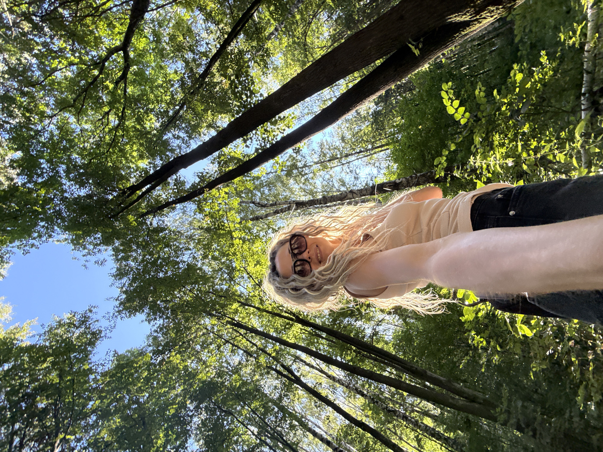 In a low-angle, wide-shot photograph taken outdoors, a woman stands in a forest. She is facing the camera, holding it out to take a selfie. The woman has long, blonde, curly hair and is wearing large, dark sunglasses, a light peach-colored sleeveless top, and black jeans. The forest setting features tall trees with green foliage and a clear blue sky visible through the canopy. The sunlight filters through the leaves, creating a dappled effect on the woman and the surrounding trees. The background is filled with trees that recede into the distance, creating a sense of depth and immersion in nature. The overall impression is one of a person enjoying a sunny day in a wooded area.