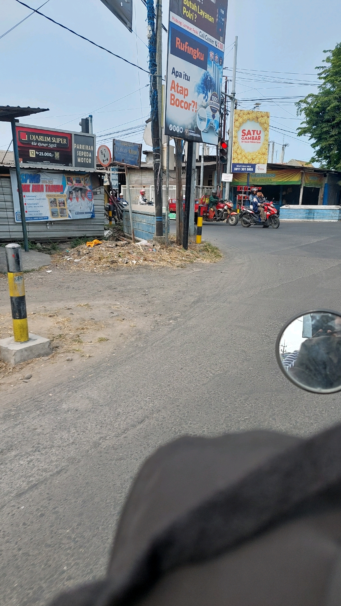 The image captures a street scene in Pilang, Indonesia, likely near a busy intersection. In the foreground, a portion of a motorcycle's handlebar and rider's torso are visible, suggesting the photo was taken from a moving vehicle. The road surface is asphalt with some gravel and debris scattered on the side.
Dominating the background are several advertising billboards and signs. One prominent billboard on the left advertises "DJARUM SUPER" with a price indication of "Rp 25.000,-" and "PARKIRAN SEPEDA MOTOR". Another sign reads "RAHMAN - RAHIM". Further to the right, a large vertical banner with the text "RUFINKU" and "Apa itu Atap Bogor?!" is displayed. Below this, a bright yellow sign features smiling faces and the text "SATU GAMBAR".
Traffic is present, with at least two motorcycles visible in motion. One red motorcycle has two riders, and another red vehicle, possibly a three-wheeled transport, is also on the road. A traffic light is illuminated red, indicating a stop.
The environment appears to be somewhat cluttered, with piles of dry vegetation and trash on the side of the road, particularly near the base of the utility poles supporting the signs. Utility poles with numerous wires are a significant feature of the scene. The sky is overcast. The presence of multiple advertisements suggests a commercial or public area within the town.