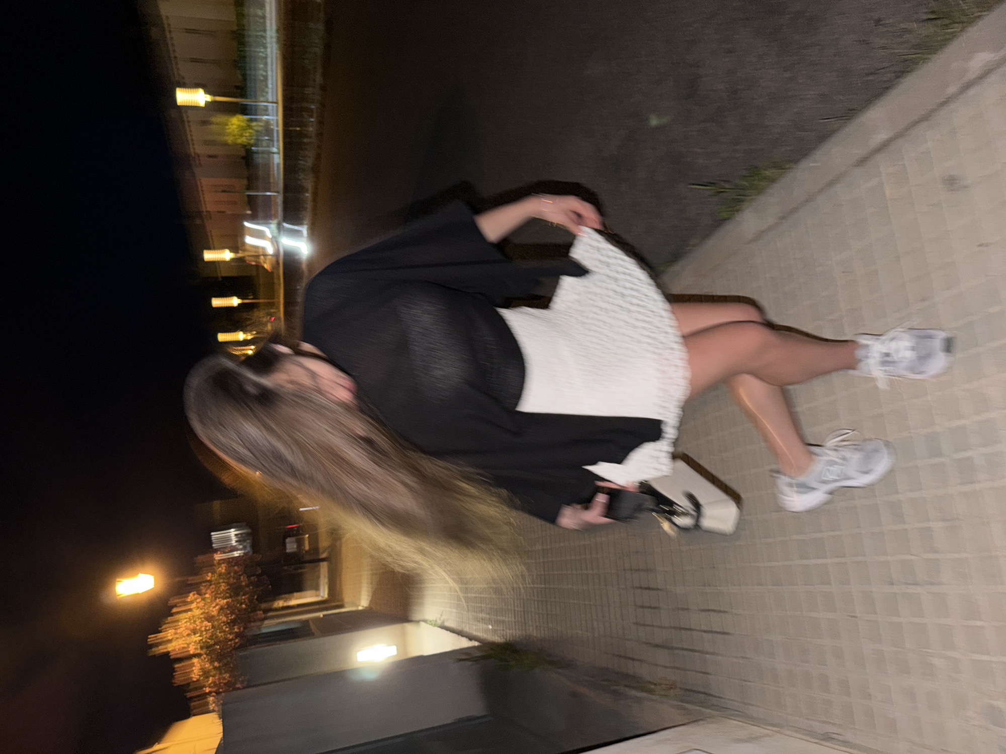 A blurry, rotated photograph shows a person sitting on a sidewalk at night. The person has long blonde hair and is wearing a black top and a white dress. They are wearing white sneakers. The sidewalk is made of gray bricks. To the left of the sidewalk is a dark area, and further back, blurred streetlights and buildings are visible. The location is possibly Platja D'alcúdia, Spain, given the information provided.