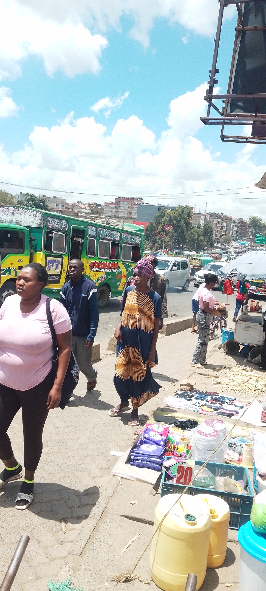 The image depicts a bustling street scene in Nairobi, Kenya. A green and yellow matatu, emblazoned with the words "FORWARD TRAVELLERS" and "PEACE MAKER", is stopped at the curb. Pedestrians, including a woman in a pink shirt and black leggings, a man in a blue jacket, and a woman in a long blue and gold dress and headwrap, are walking along the sidewalk.
A vendor's display is set up on the sidewalk with various items for sale, including packages of sanitary pads, socks, party hats, and yellow jerrycans with the number "20" displayed. In the background, there are other vehicles and buildings visible, with a partly cloudy sky overhead.