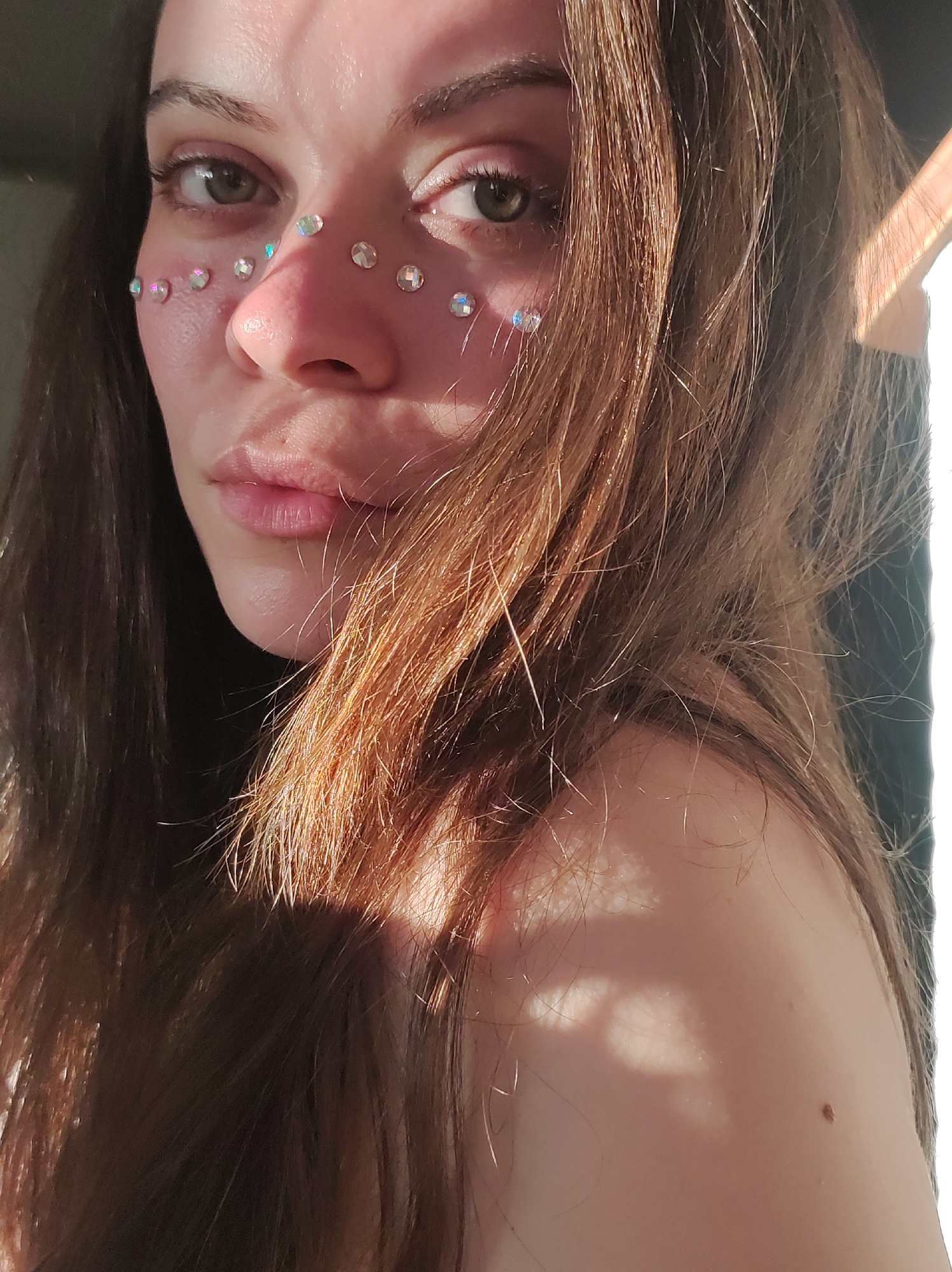A young woman with long brown hair is the focus of the image. She has fair skin, brown eyes, and is wearing iridescent rhinestones arranged from the inner corners of her eyes, down her cheeks, and across the bridge of her nose. Sunlight shines on her face and shoulder, and casts shadows on the right side of the image. The setting is unclear, but based on the request, it is theoretically in the city of Фоки, Russia. However, there is no visual information in the image that confirms this location.