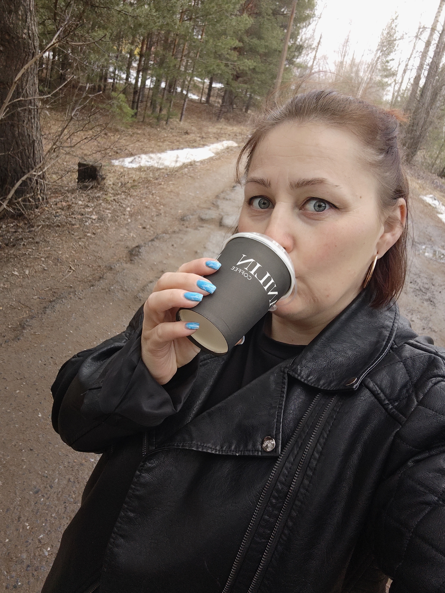 A woman with fair skin and blue eyes is drinking from a black coffee cup in an outdoor environment. The woman is wearing a black leather jacket and has blue nail polish. She is positioned on a dirt road with trees and some snow in the background. The coffee cup has white writing on it. The location appears to be a wooded area in Chaikovsky, Russia.