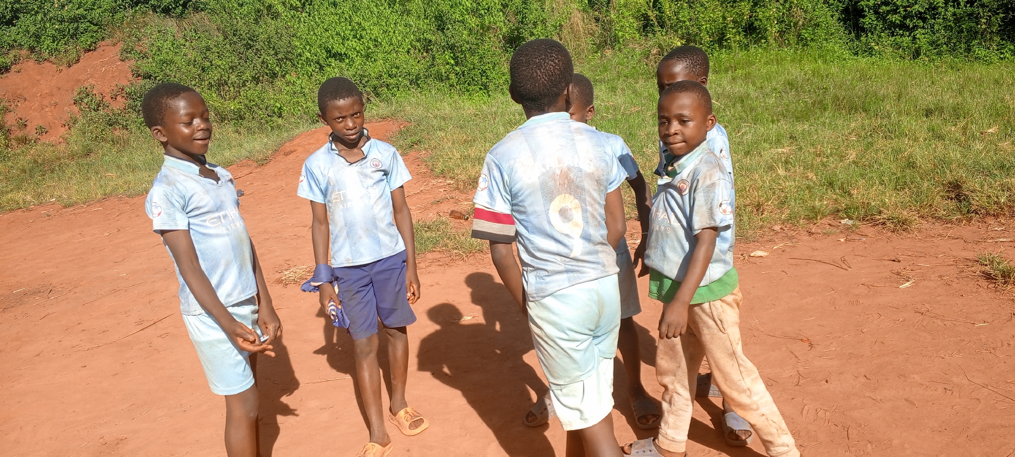 In Ngalula, Democratic Republic of the Congo, a group of five young boys are gathered on a dirt path near greenery. They are wearing matching light blue soccer jerseys with "City Airways" logos. Their shorts vary in color and style, and some wear sandals. One boy has a red and black armband. The background consists of lush green bushes and grass. The ground is red dirt.