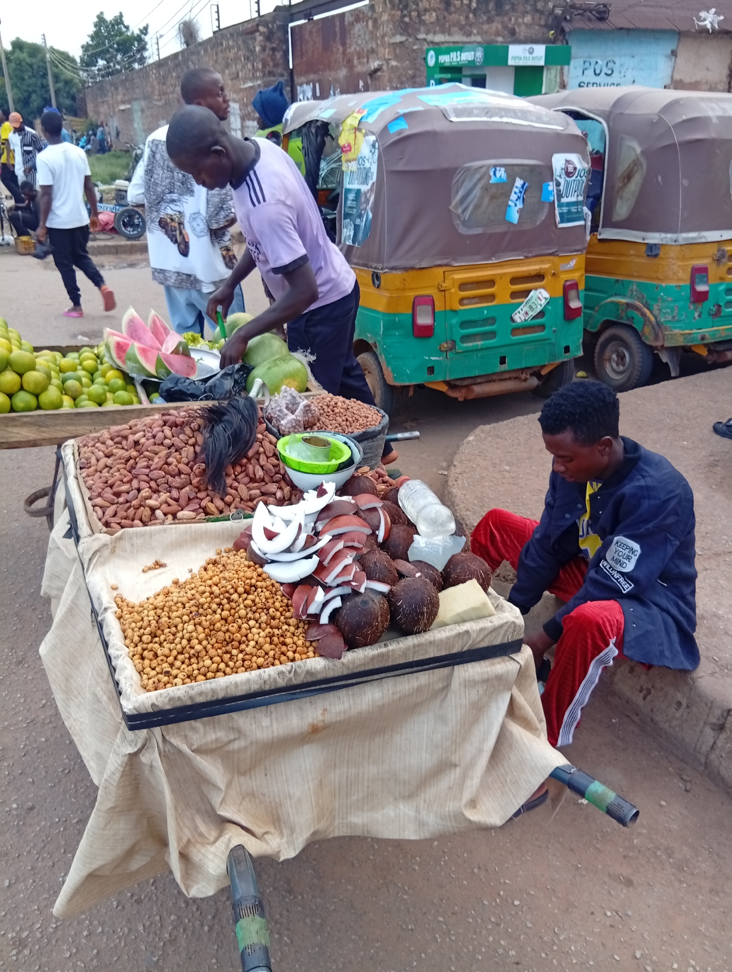 In Bukuru, Nigeria, a street vendor prepares to sell goods from a cart. The cart is laden with a variety of items, including a large quantity of what appear to be dried beans or nuts in a wooden crate. Sliced coconuts are also visible, along with whole coconuts and what might be a block of cheese or butter. To the left, crates of oranges and other citrus fruits are stacked. In the background, three auto-rickshaws, common in Nigerian cities, are parked. One is predominantly yellow and green, another is green and yellow, and the third is mostly green. The street is paved with asphalt and dirt. A man in a blue jacket and red trousers sits beside the cart, attending to his wares, while another man in a purple shirt is seen interacting with the fruits on the cart. Several other individuals are present in the background, engaged in their daily activities. The overall scene captures a bustling moment of commerce on a Nigerian street.