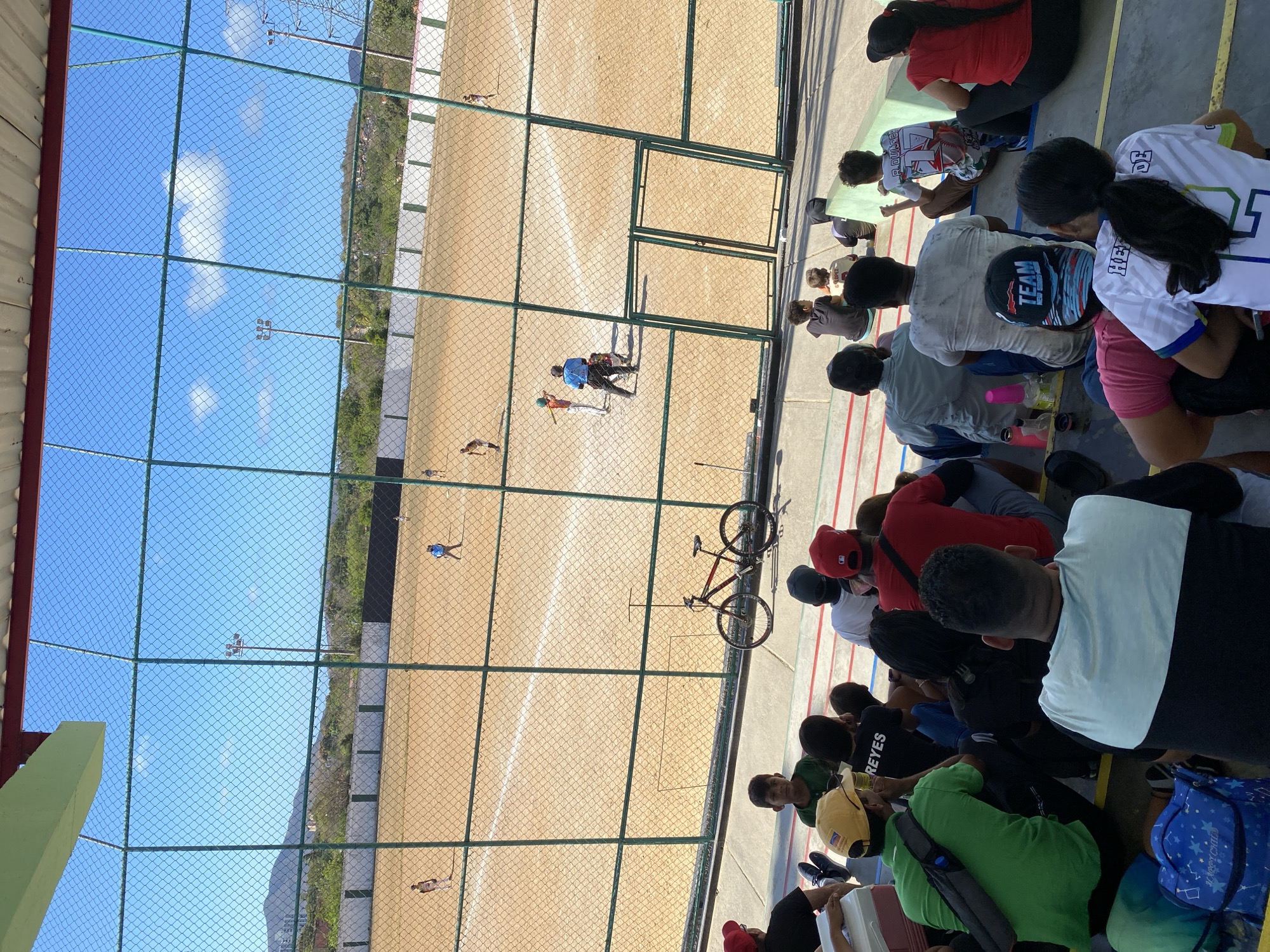 The image depicts a baseball field in Juan Griego, Venezuela, viewed from behind a chain-link fence, likely from stadium seating. People are seated in rows, watching a baseball game. A bicycle is resting against the fence, seemingly unattended. The field itself is a light tan color with white baselines. Several players are visible on the field, involved in a game. Beyond the field, trees and possibly a hillside are visible. The sky is blue with some clouds.