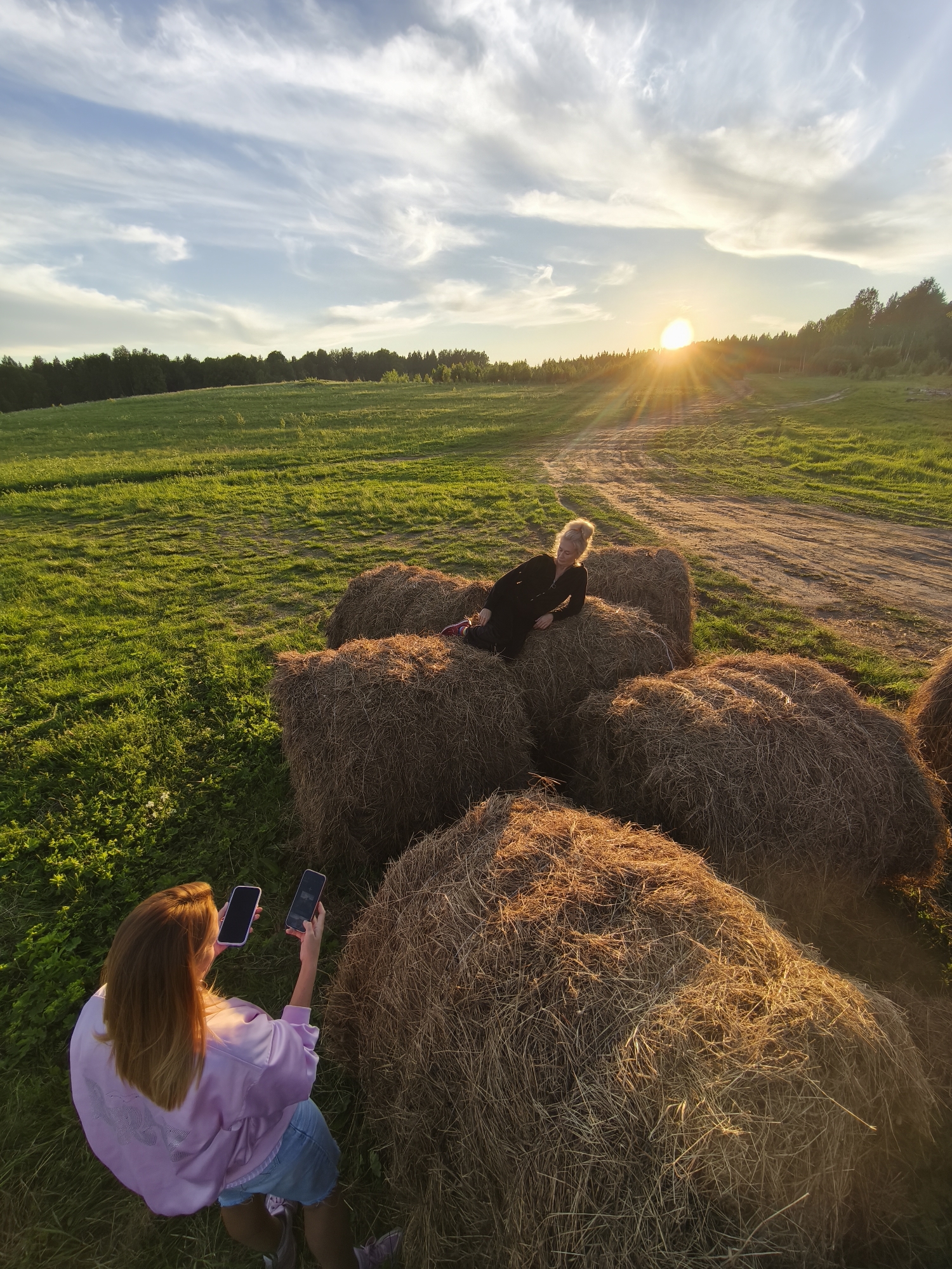 The image depicts an outdoor scene at sunset in a rural landscape, likely Стеклянный, Russia. In the mid-ground, a woman with blonde hair, dressed in a black long-sleeved top and dark pants, is posing on a stack of large, round hay bales. She reclines on one bale while another woman, positioned in the foreground with her back to the viewer, is actively photographing her. This photographer has long brown hair and is wearing a light pink, possibly satin, long-sleeved top and denim shorts. She holds two smartphones, both with dark screens, up towards the subject.
The setting is an expansive green field, illuminated by the warm, golden light of the setting sun. The sun is visible on the horizon, partially obscured by a line of dark green trees that stretch across the background. A dirt path or road winds through the field towards the distant tree line. The sky above is a blend of light blue and soft white, with wispy cirrus clouds adding texture. The hay bales are bathed in the golden hour light, highlighting their straw textures. The overall atmosphere is serene and vibrant, capturing a moment of photography amidst the beauty of the countryside during dusk. No visible text is present in the image.