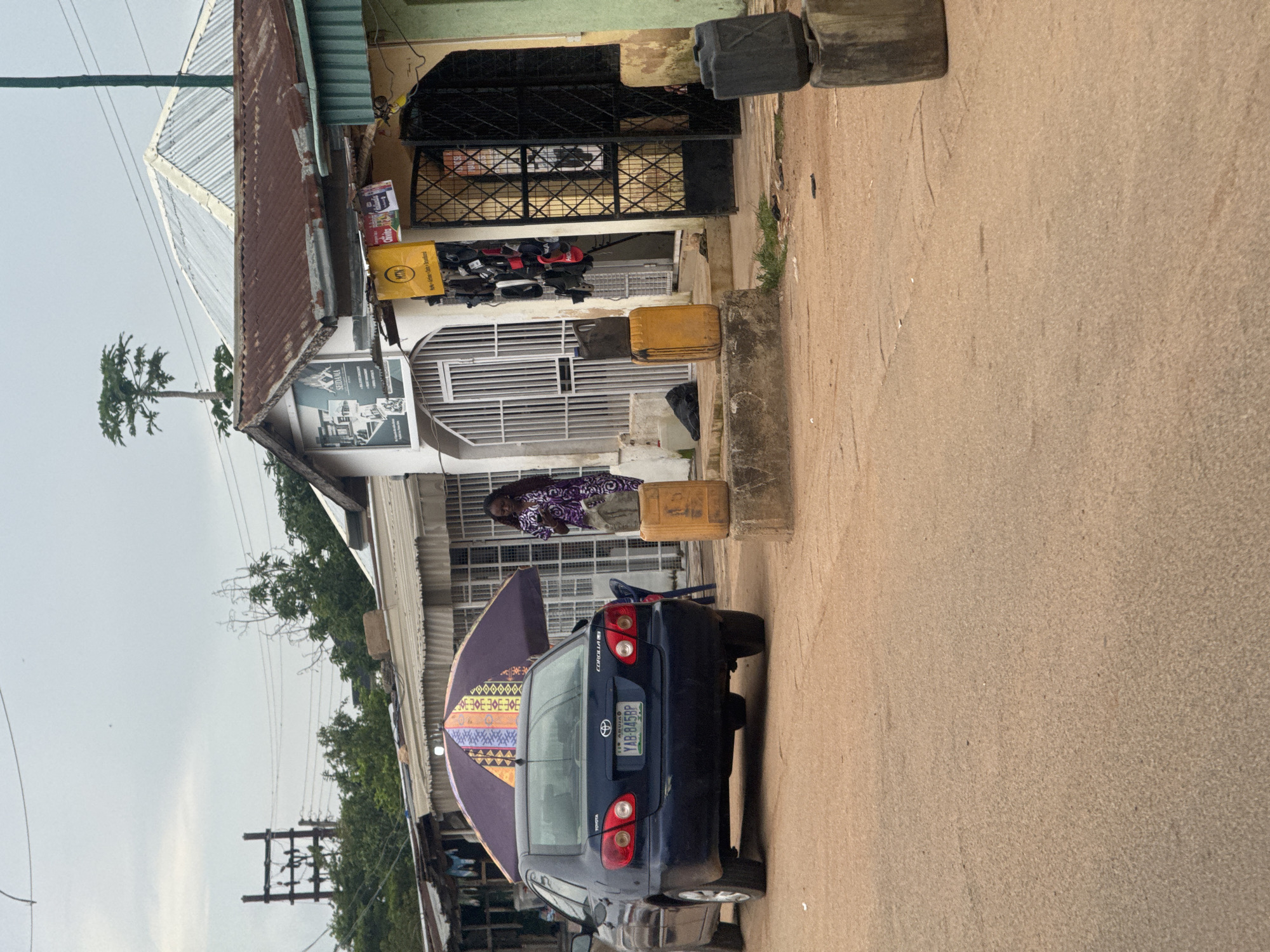 In Gura Top, Nigeria, a dark blue Toyota Corolla is parked on a dirt road in front of several small shops. The car's license plate reads YAB-845BP. To the right of the car, a woman in a purple patterned dress stands next to a yellow jerrycan. A large, patterned umbrella is open behind the car, providing shade. The shops have corrugated metal roofs and open doorways. One shop displays various items hanging outside, including what appear to be shoes and bags. Yellow and black jerrycans are stacked on concrete blocks in front of the shops. In the background, utility poles and wires are visible, along with lush green trees. The sky is overcast.